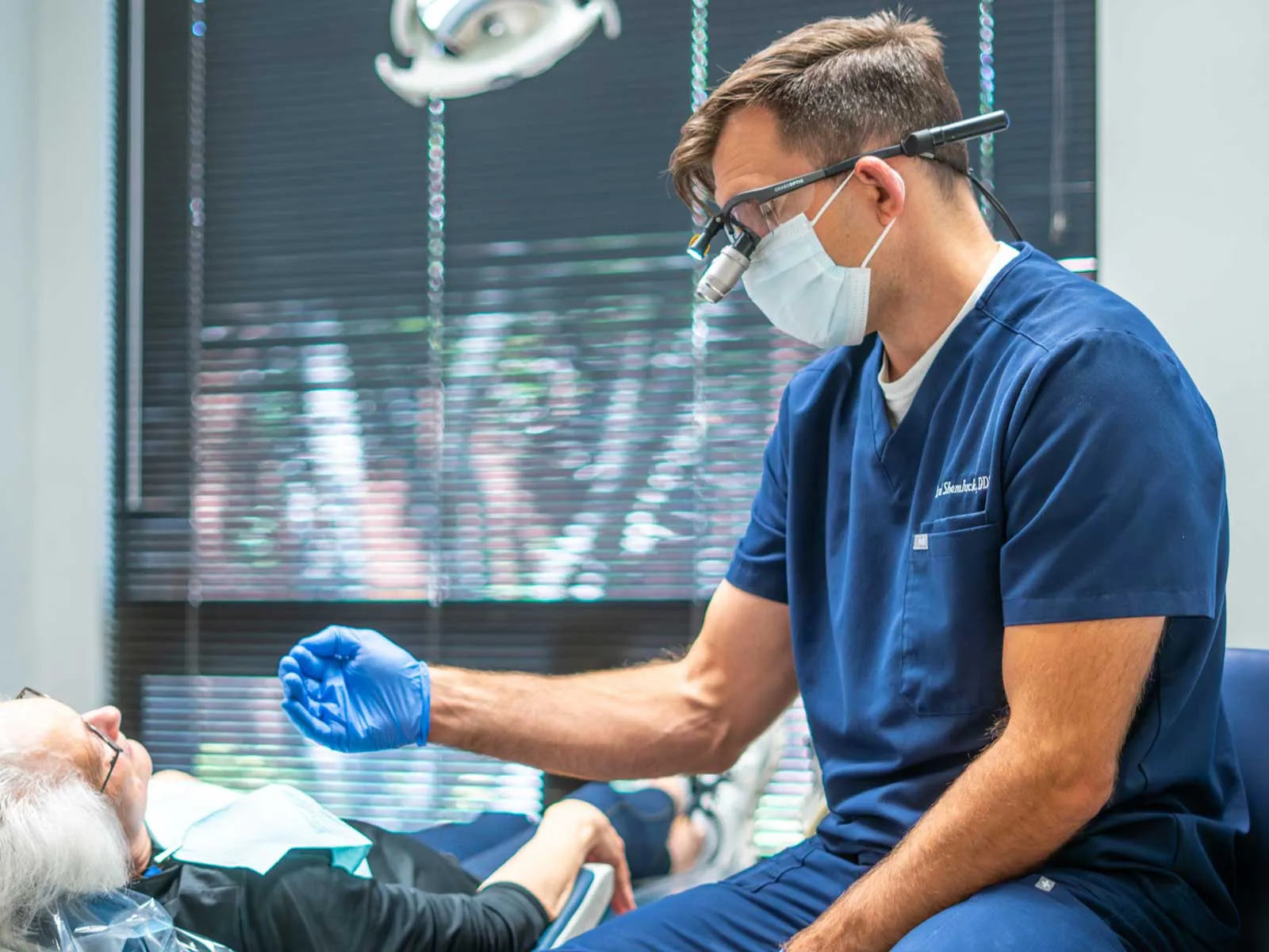 Dentist in blue scrubs and mask examining elderly female patient in dental clinic with natural light.