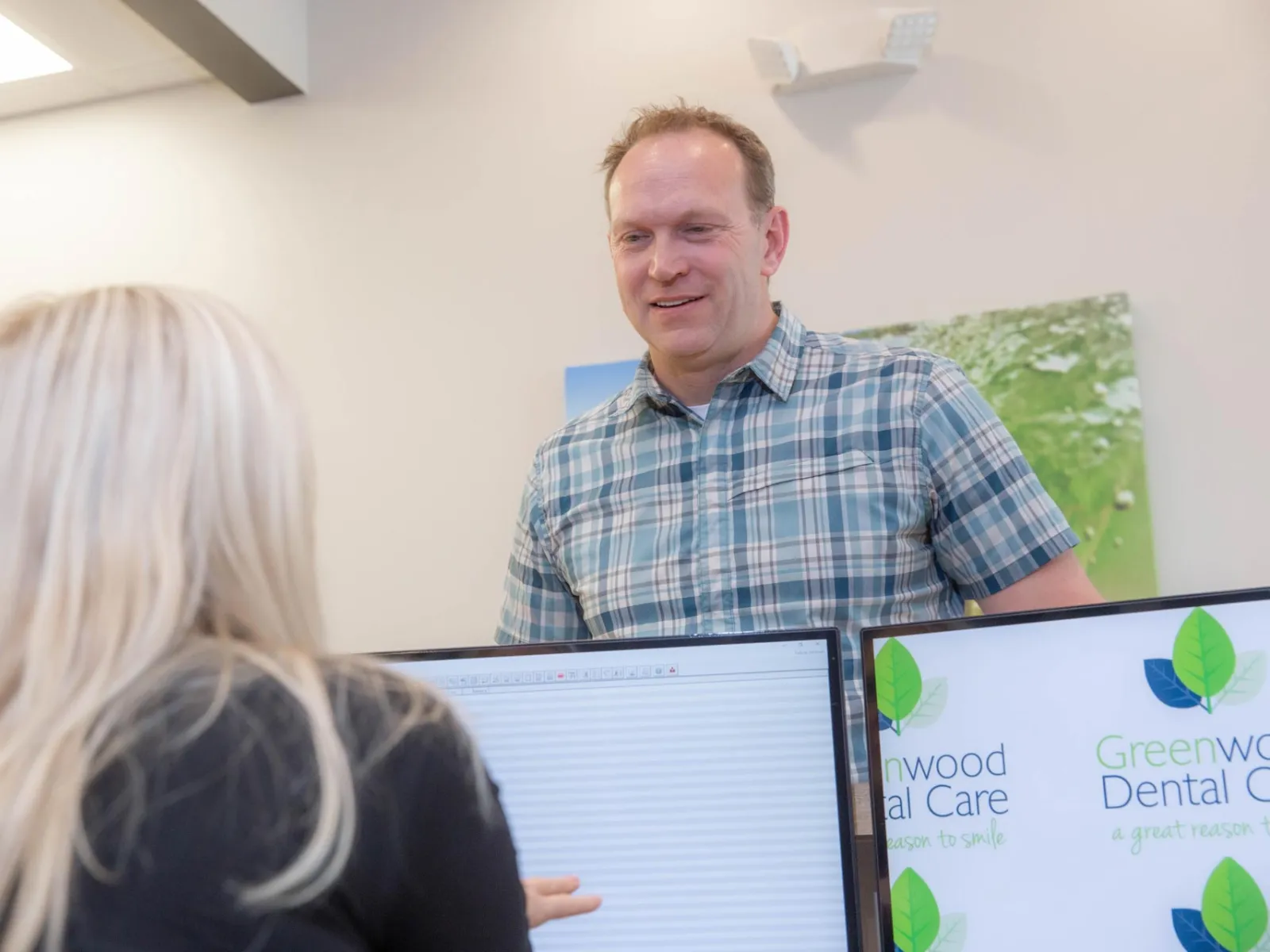Man in plaid shirt speaking with woman at dental office reception with Greenwood Dental Care logo visible