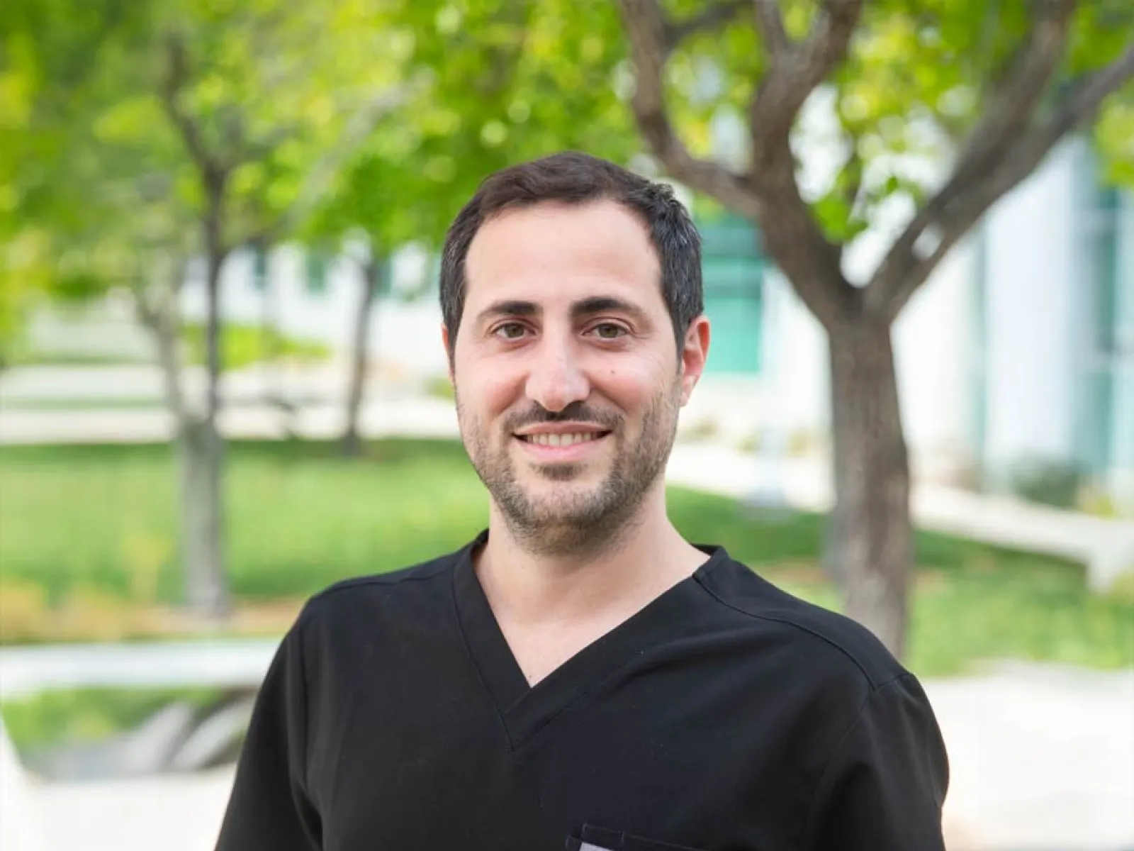Smiling healthcare professional in scrubs standing outdoors in a green setting.
