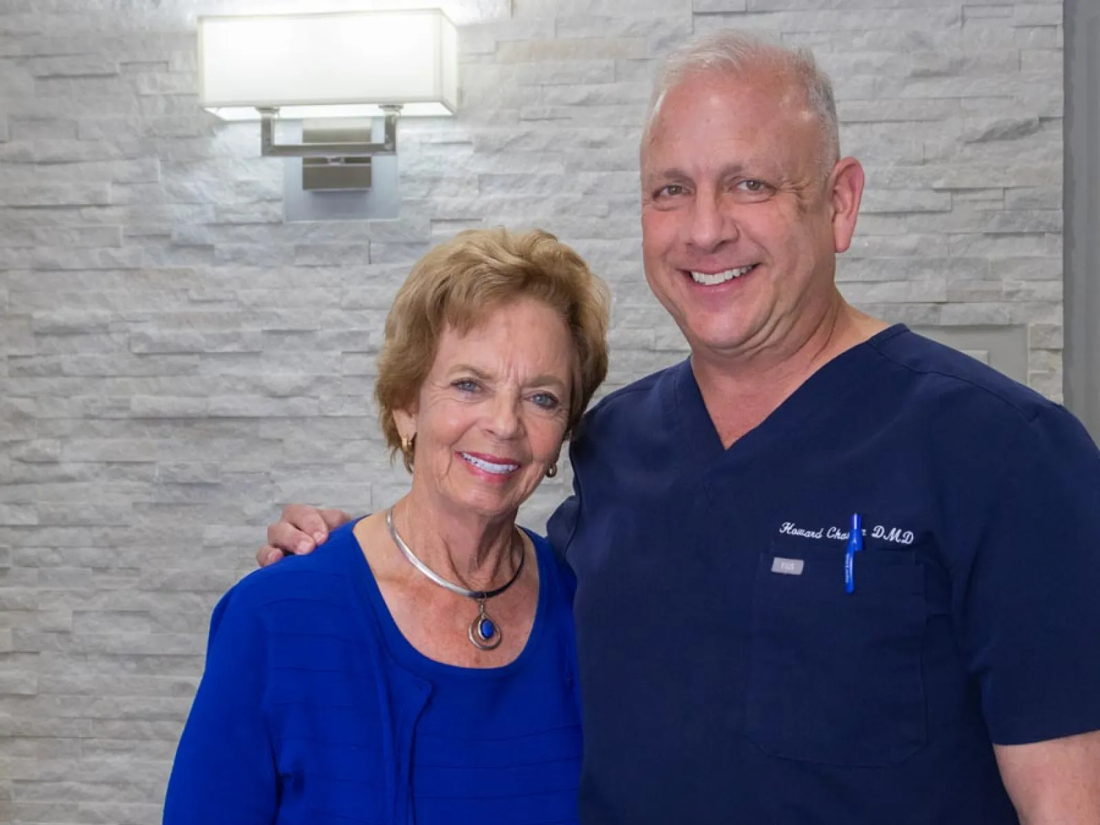 Smiling senior woman with doctor in blue scrubs standing together inside a medical office with stone wall.