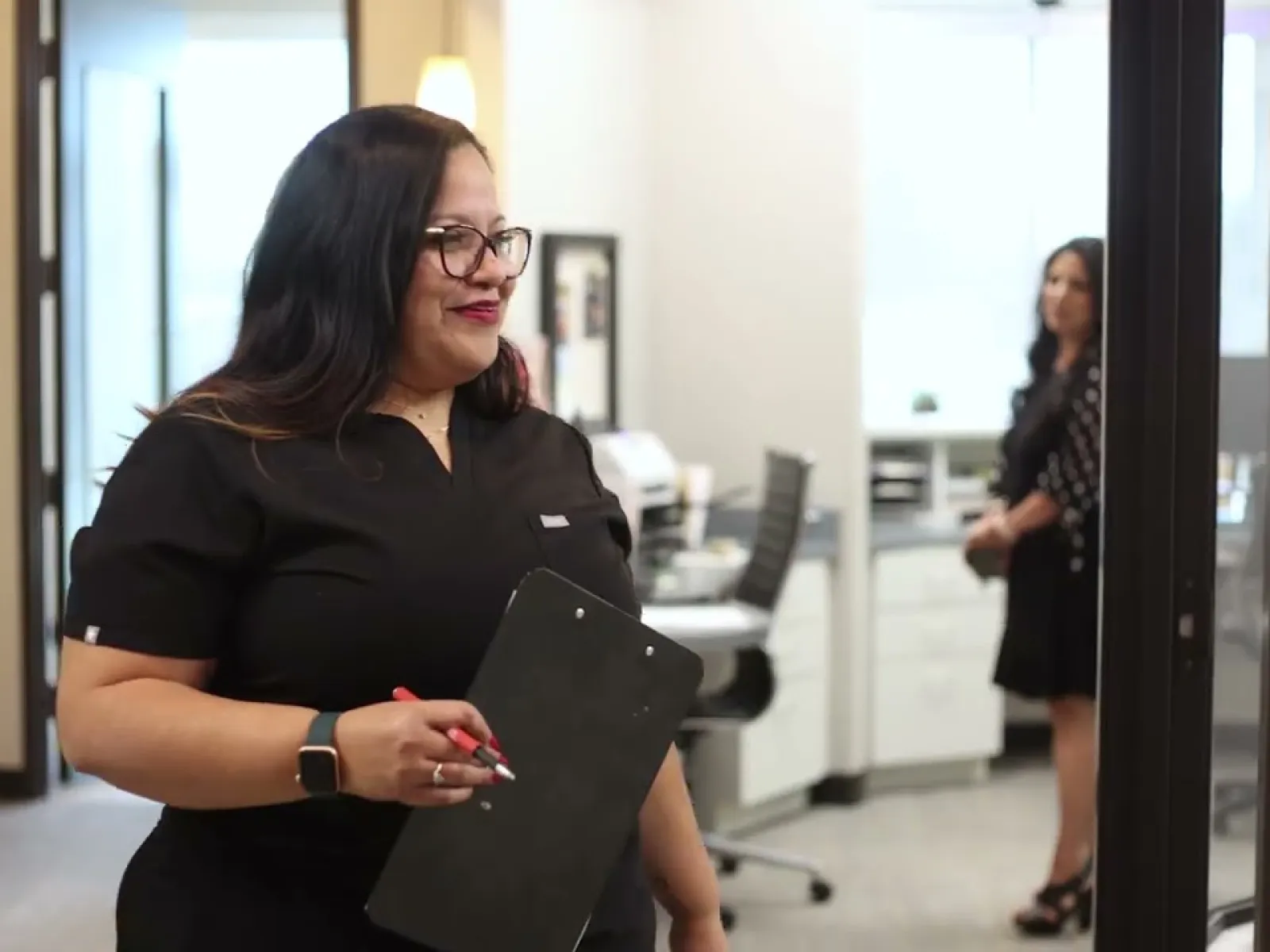 Woman in black scrubs holding clipboard walking in a modern medical office with another woman in background.