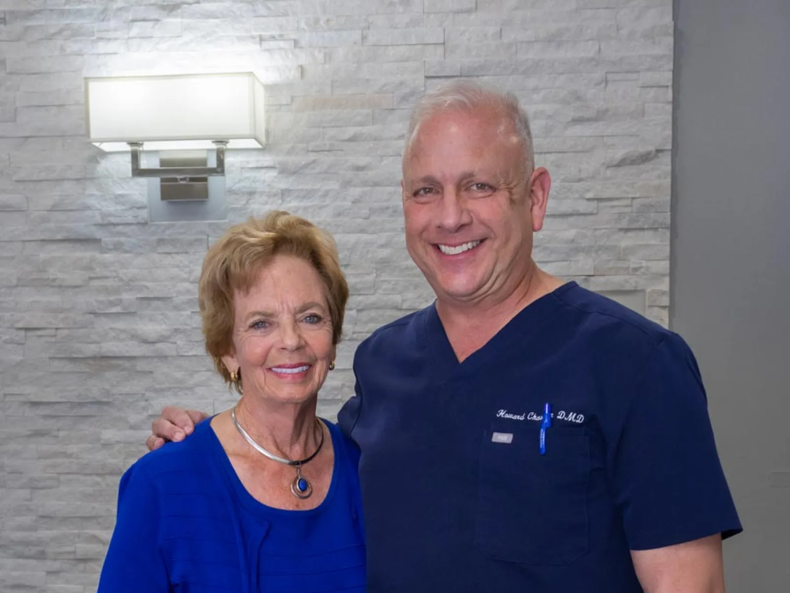 Smiling elderly woman with a man in navy blue medical scrubs in front of a stone wall and light fixture