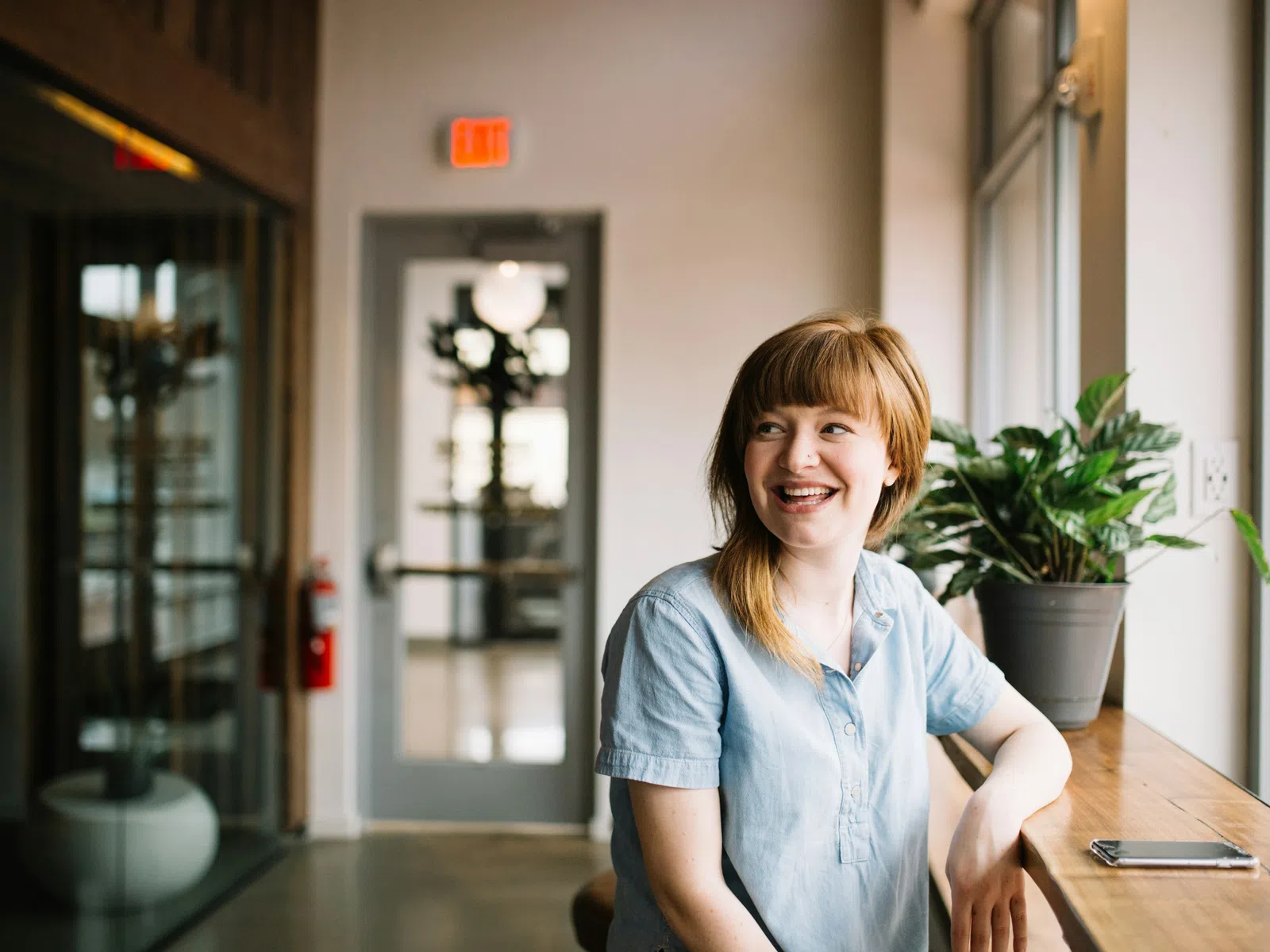 Smiling young woman sitting by a window in a bright modern cafe with plants and smartphone on the table.