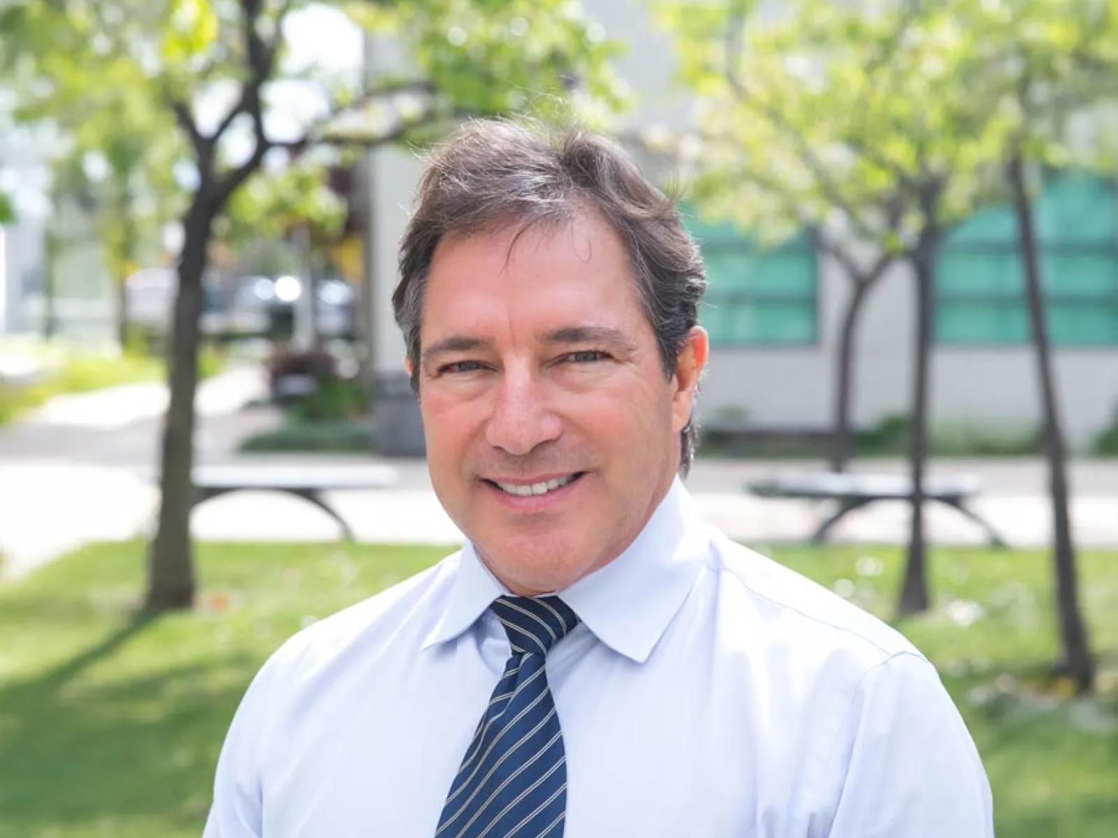 Smiling man in a dress shirt and tie standing outdoors with greenery in the background.