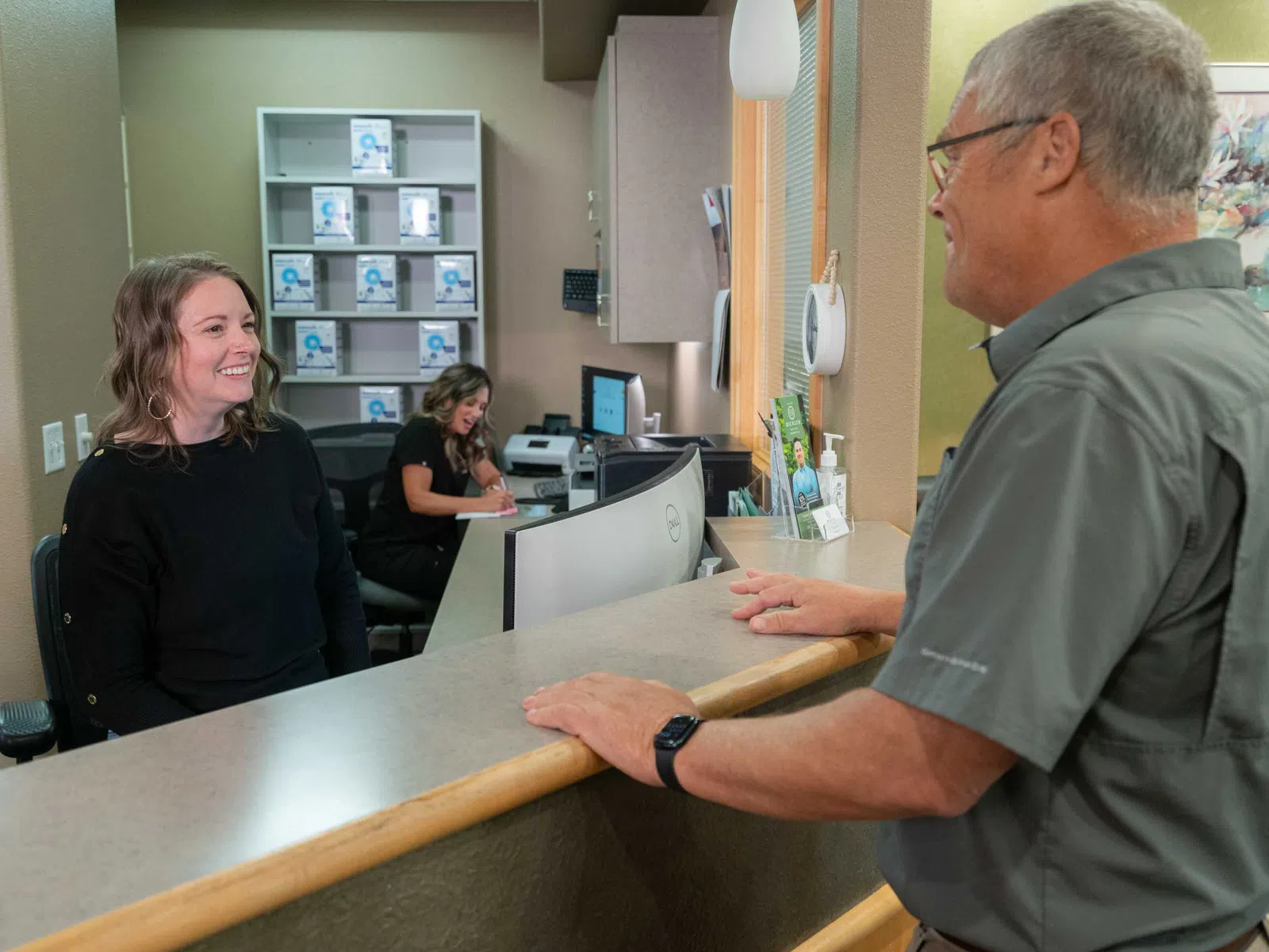 Smiling receptionist assisting an older man at a medical office front desk with another staff member working in the background