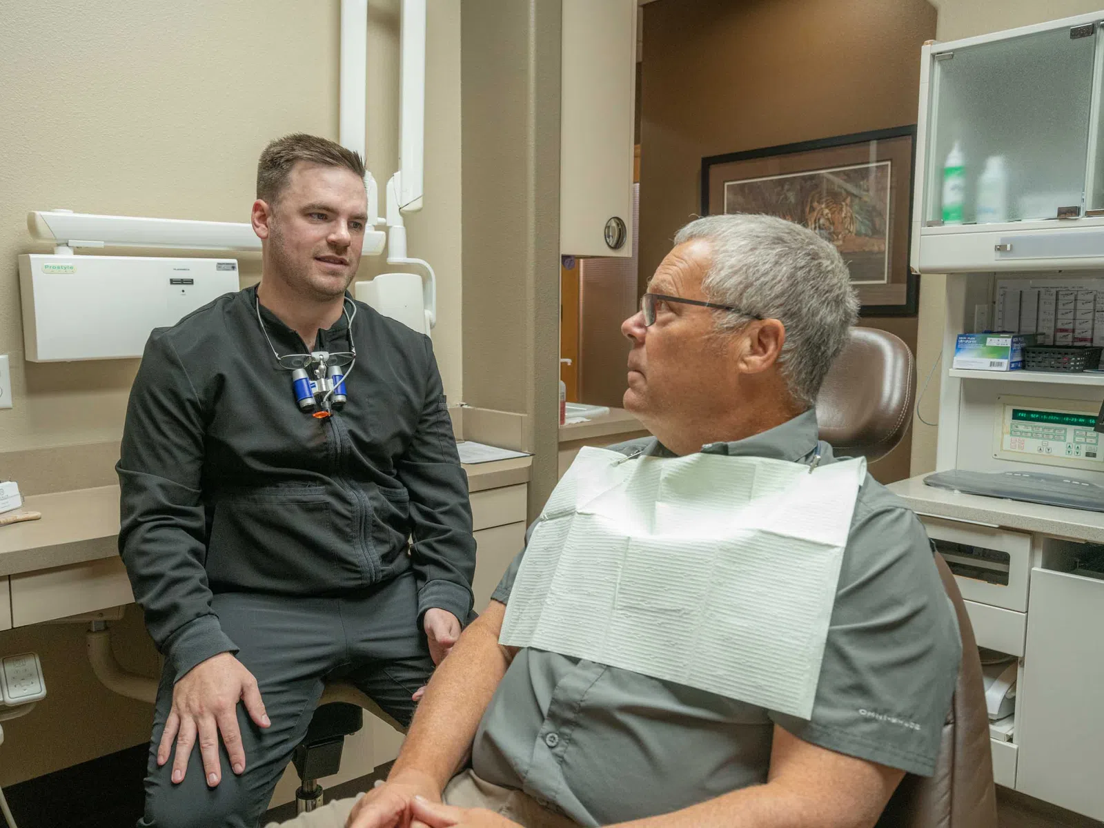 Dentist in black coat consulting older male patient seated in dental chair with bib in modern clinic room.
