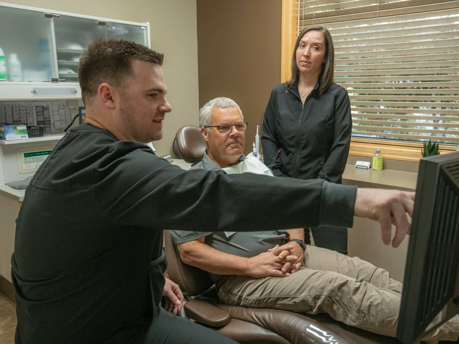 Dentist explains treatment to older male patient with female assistant observing in clinic room