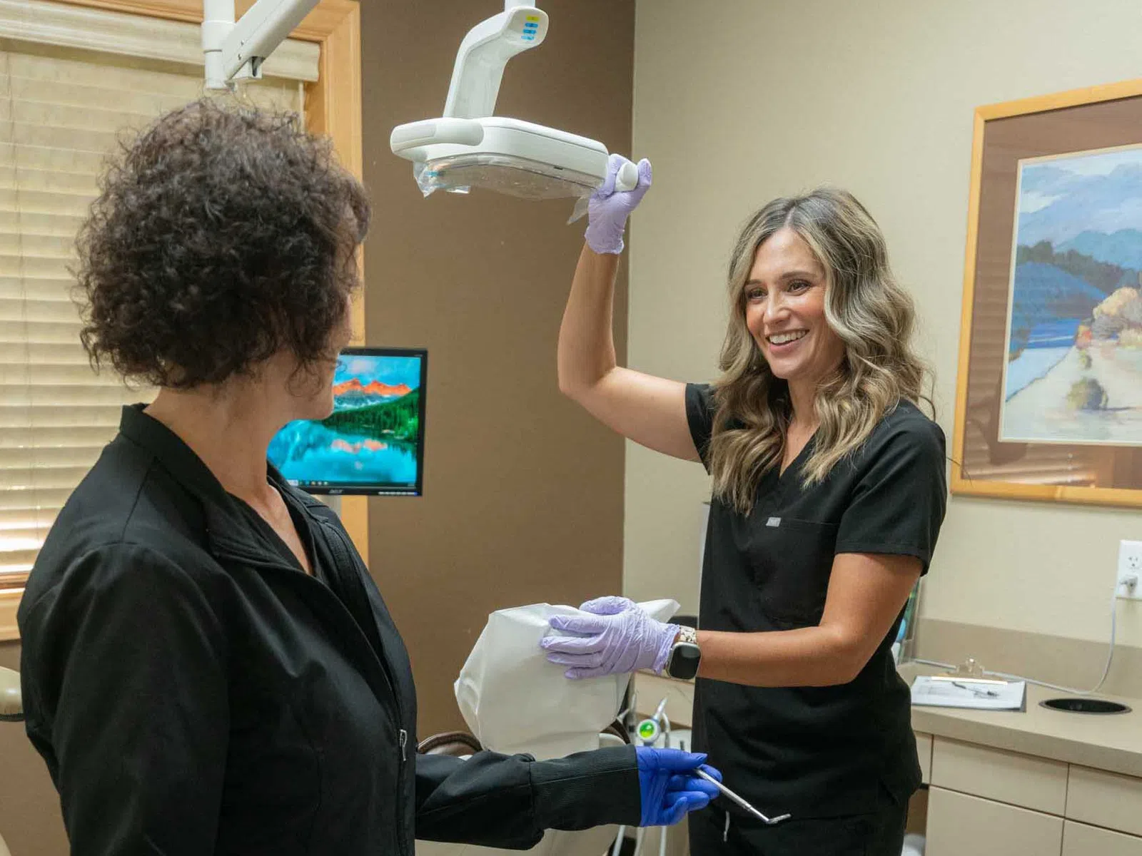Female dental professional prepares dental x-ray machine while patient sits in exam chair in clinic room.