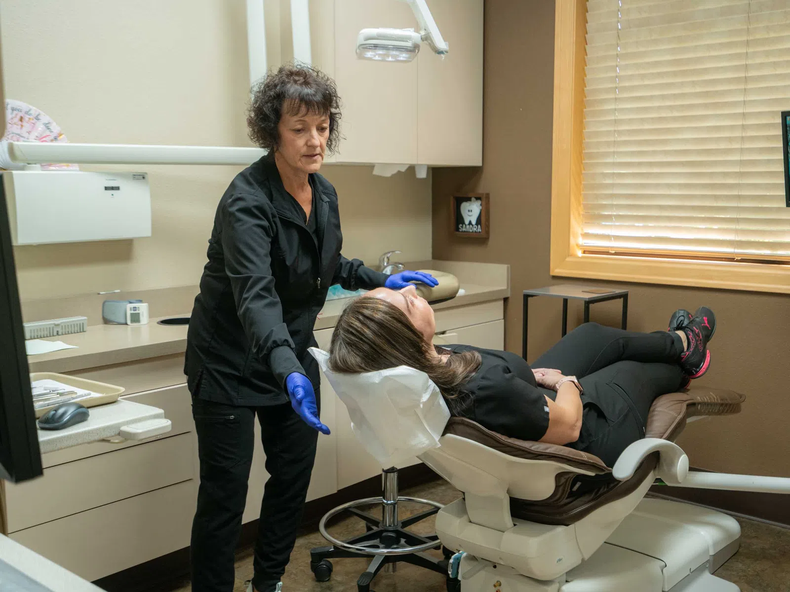 Dental hygienist adjusting reclining chair for female patient in a modern dental office with monitor.