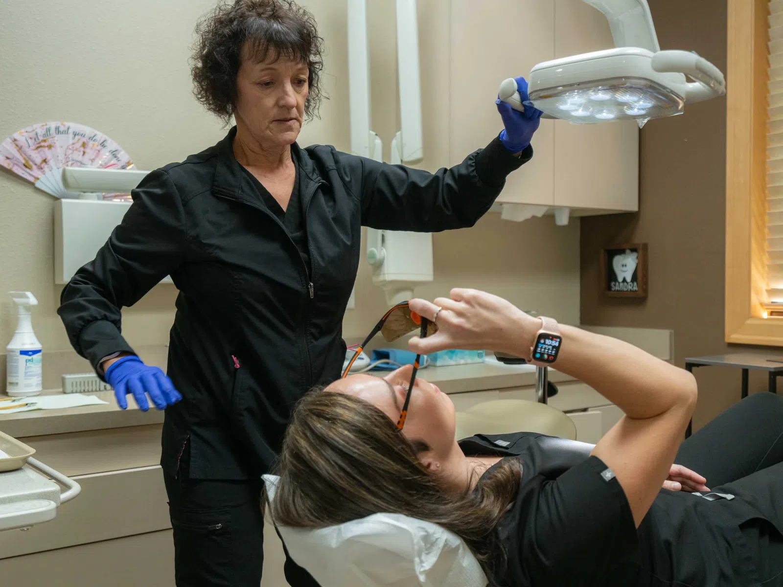Dental hygienist adjusting overhead light while patient lies down wearing protective glasses in a clinic.