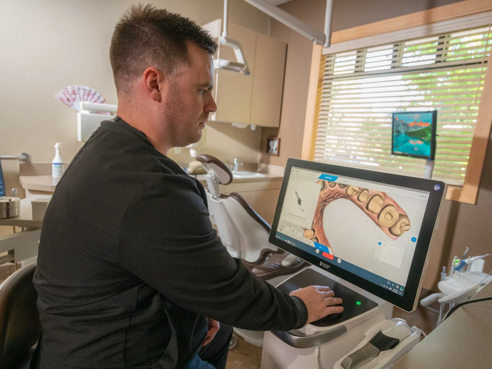 Dentist using 3D dental imaging software to examine a digital model of teeth in a clinic room.