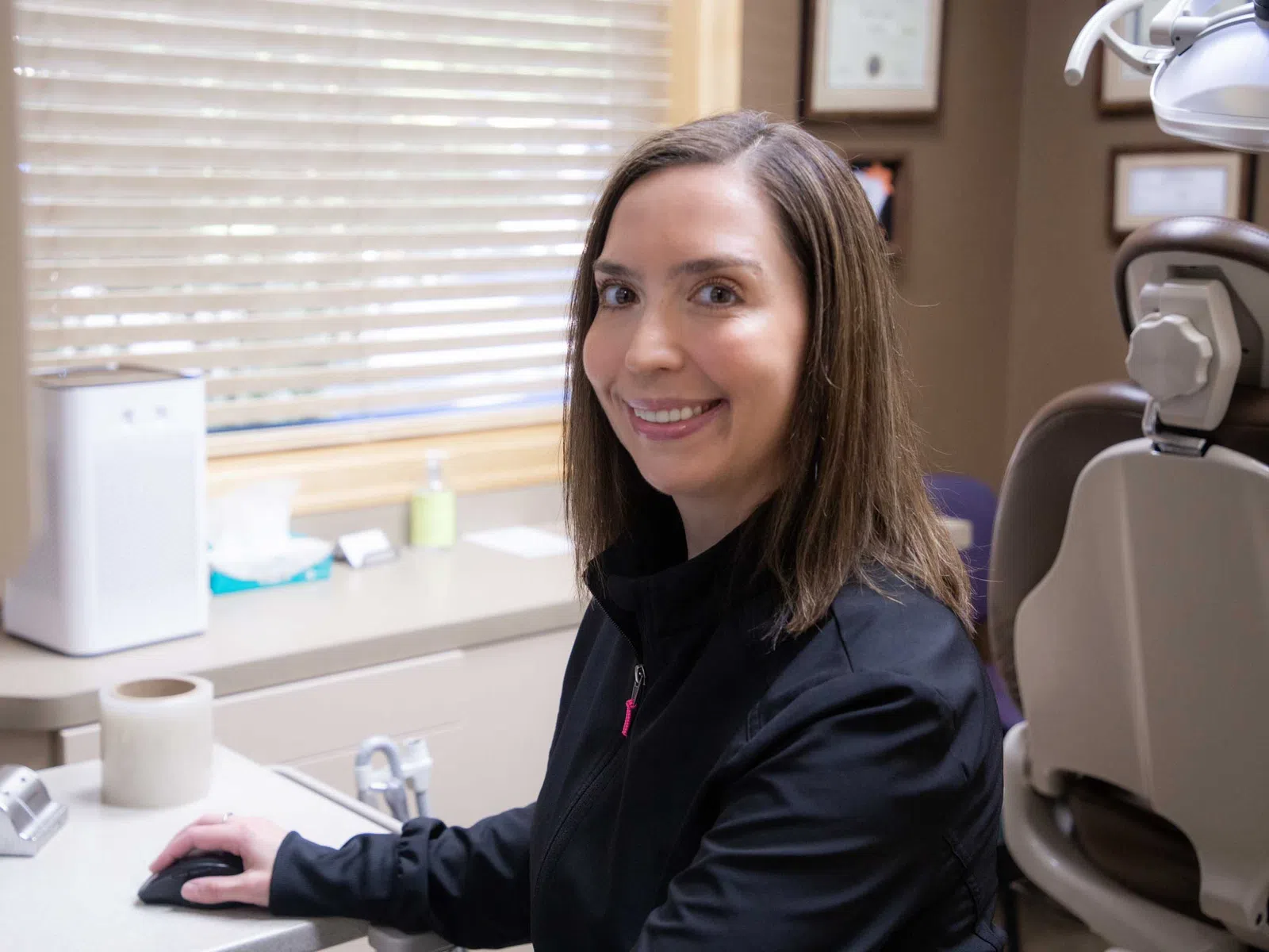 Smiling female dental professional at workstation in a bright dental office with equipment and window blinds.