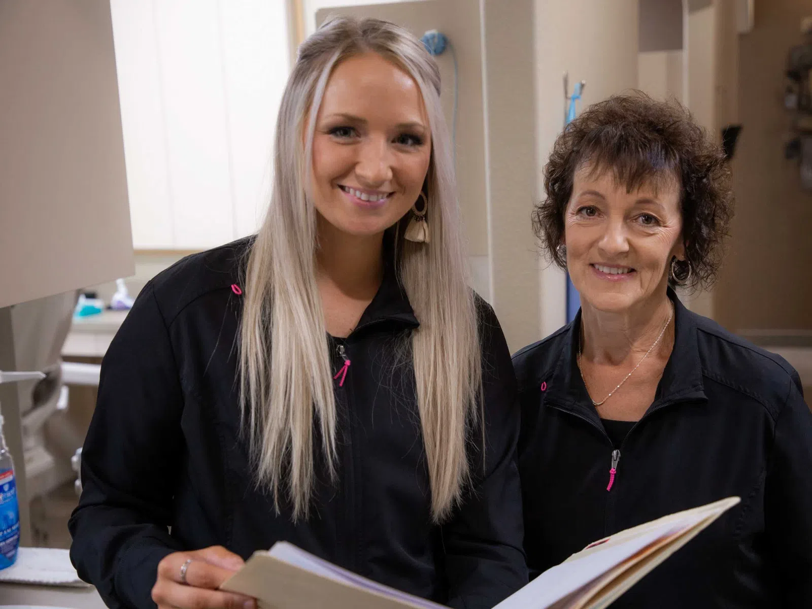 Two female healthcare workers smiling, wearing black uniforms, holding patient files in a medical office.