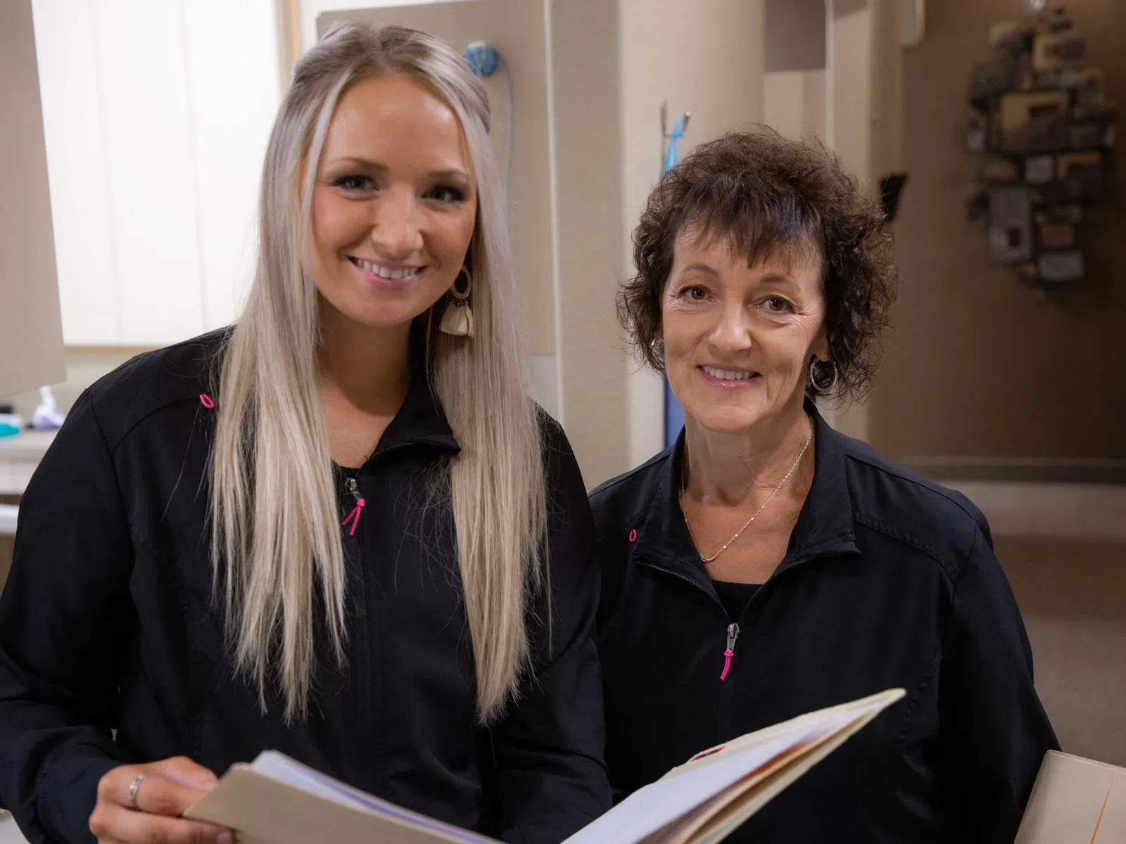 Two smiling women in black jackets holding an open folder indoors, suggesting teamwork or consultation.