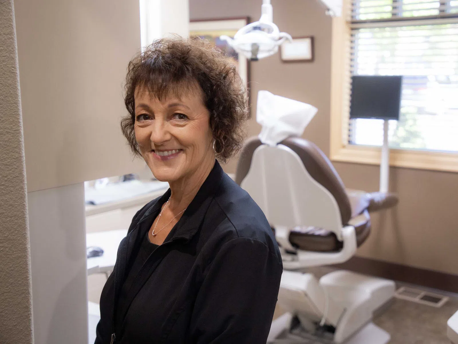 Smiling female dentist standing in dental office with chair and equipment in the background under natural light.