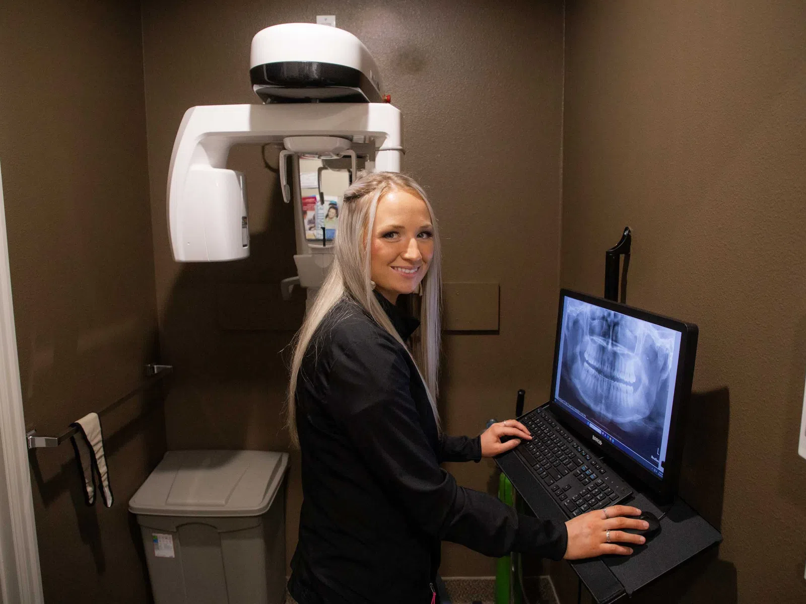 Dentist analyzing a dental X-ray on a computer in a clinic next to an X-ray machine, smiling and ready to assist.