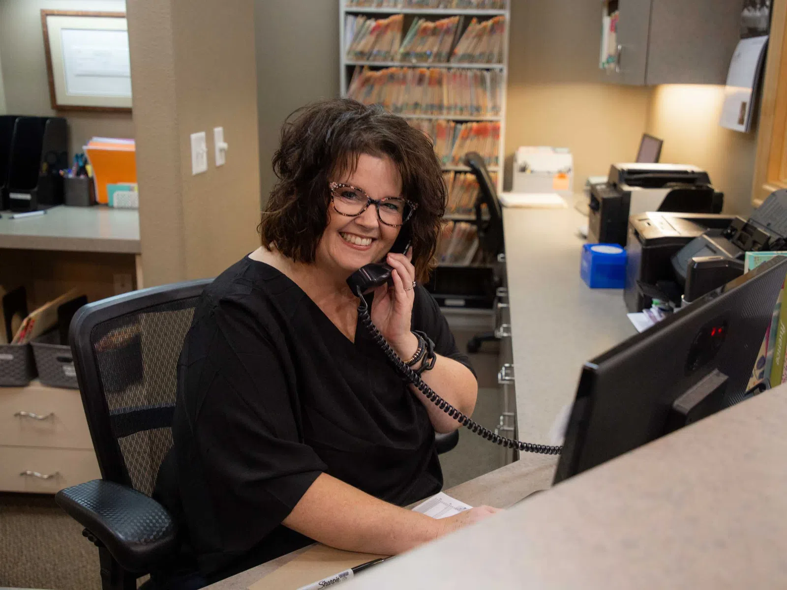 Smiling woman receptionist talking on phone at a busy office desk with files and computer.