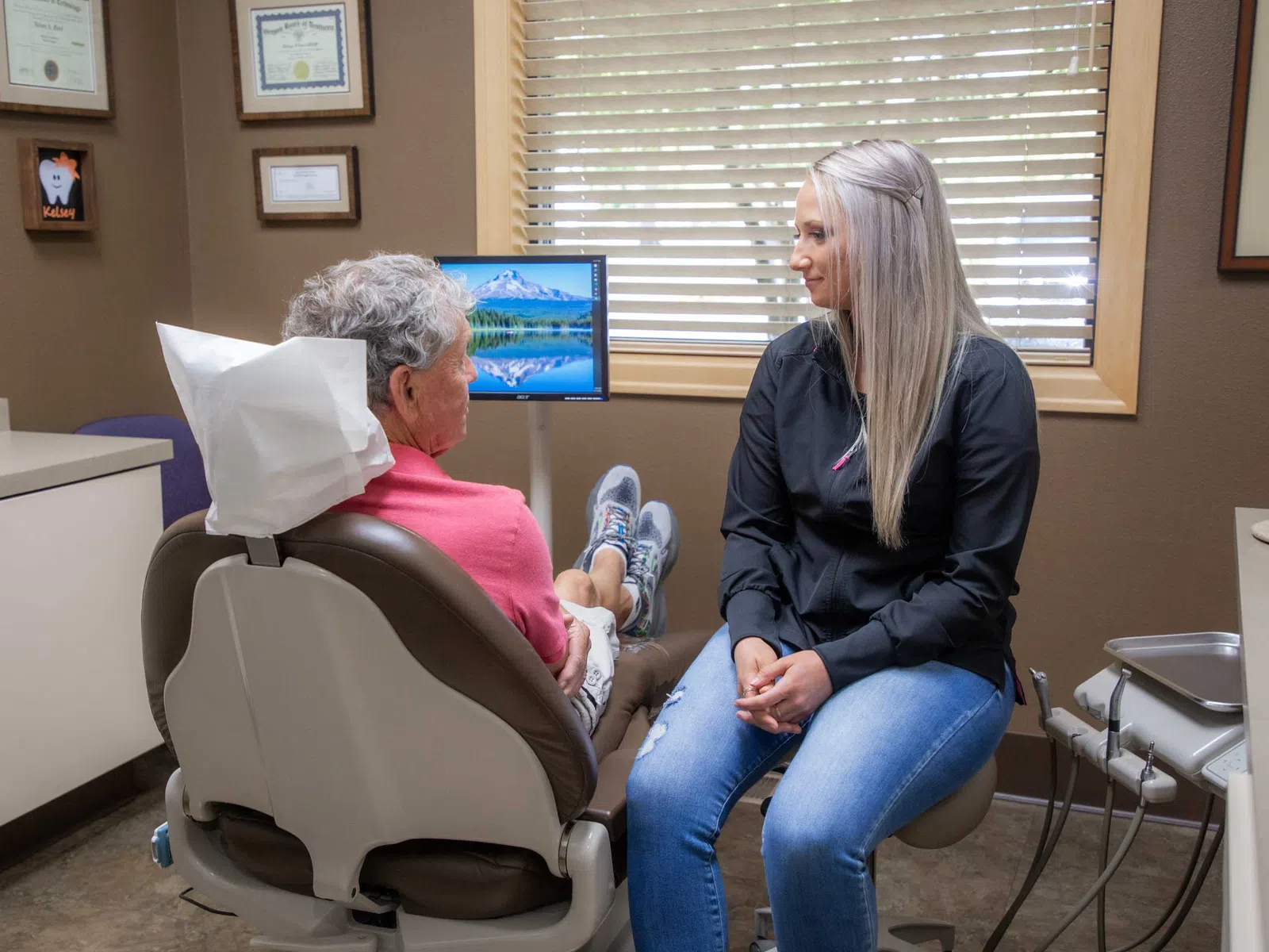 Dentist or hygienist talking to patient in dental office with window blinds and computer screen.