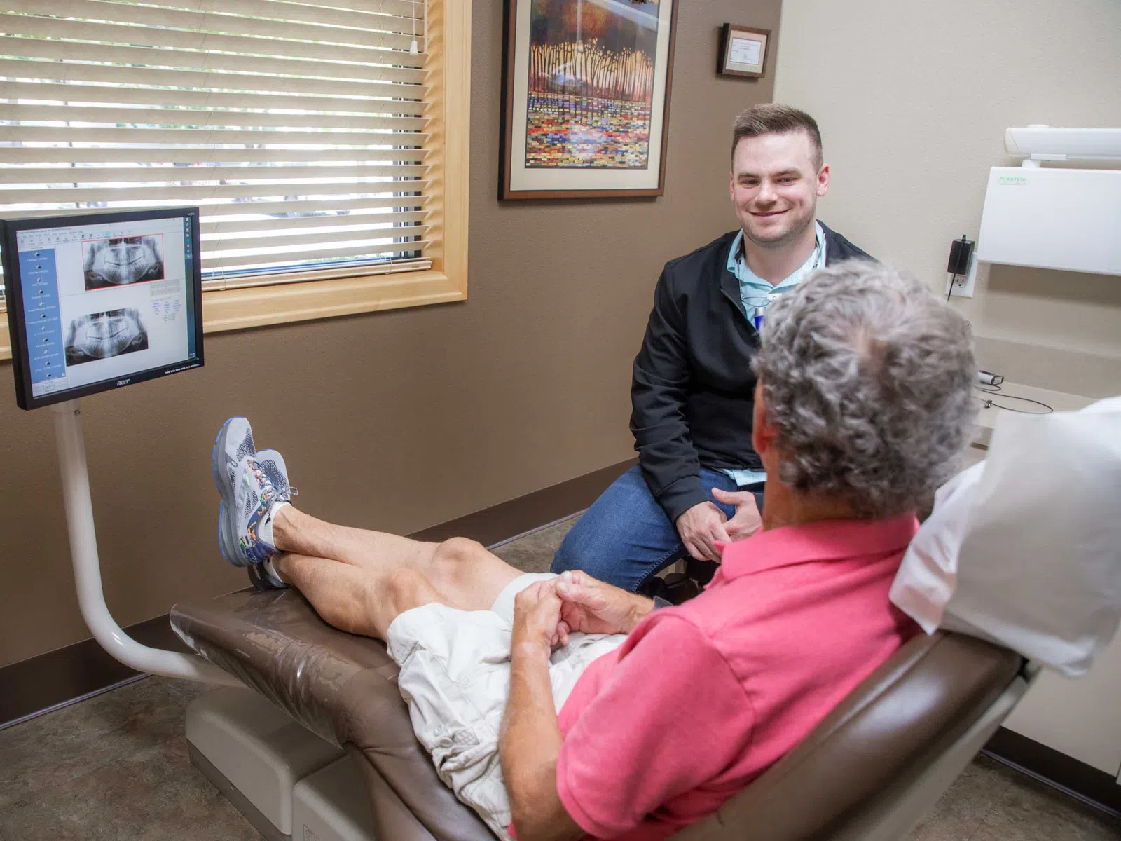 Dentist consulting with patient in exam room with dental X-ray images displayed on monitor.