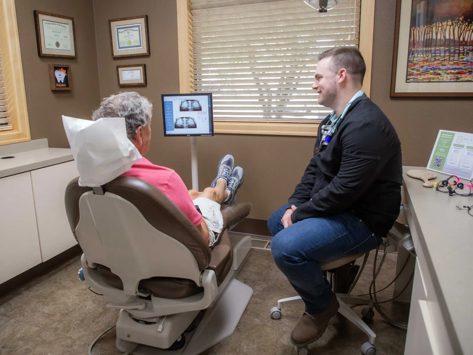 Dentist consulting with elderly patient in dental office with X-ray images on a monitor and certificates on the wall