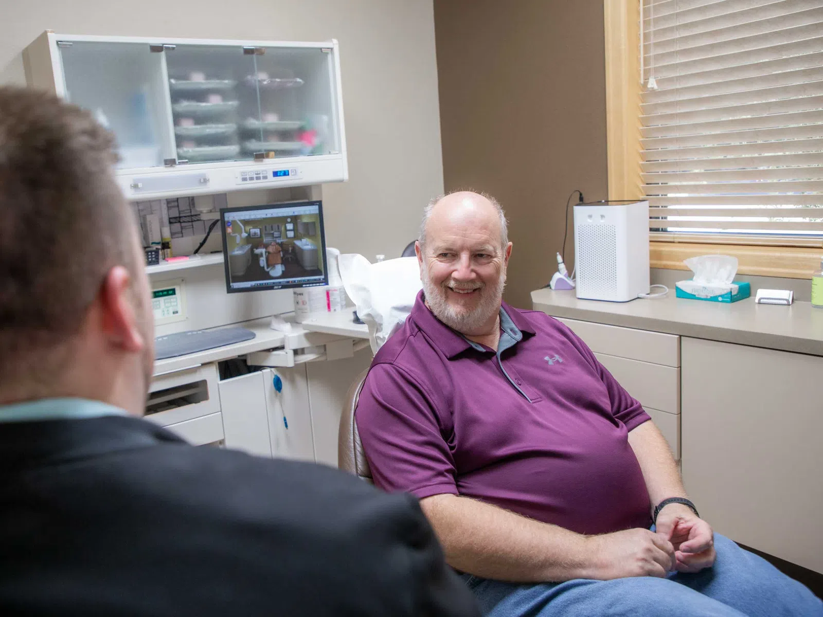 Older man in purple shirt smiling and talking to a doctor in a medical office with medical equipment.