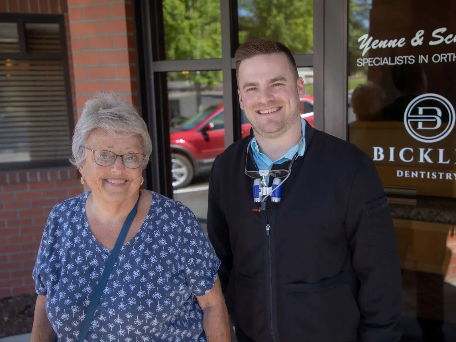 Smiling elderly woman and male dentist standing outside Bickler Dentistry office with glass door and signage.