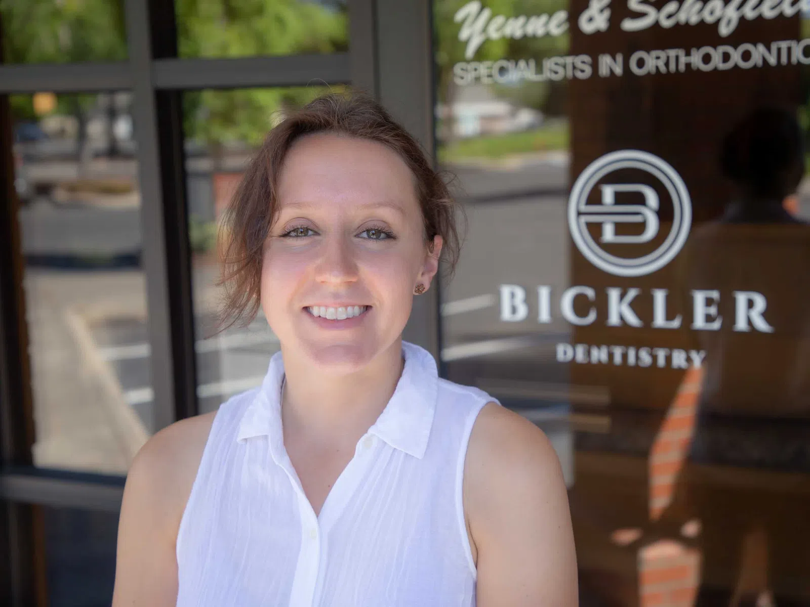 Smiling woman in white sleeveless blouse standing in front of Bickler Dentistry and orthodontics office door.