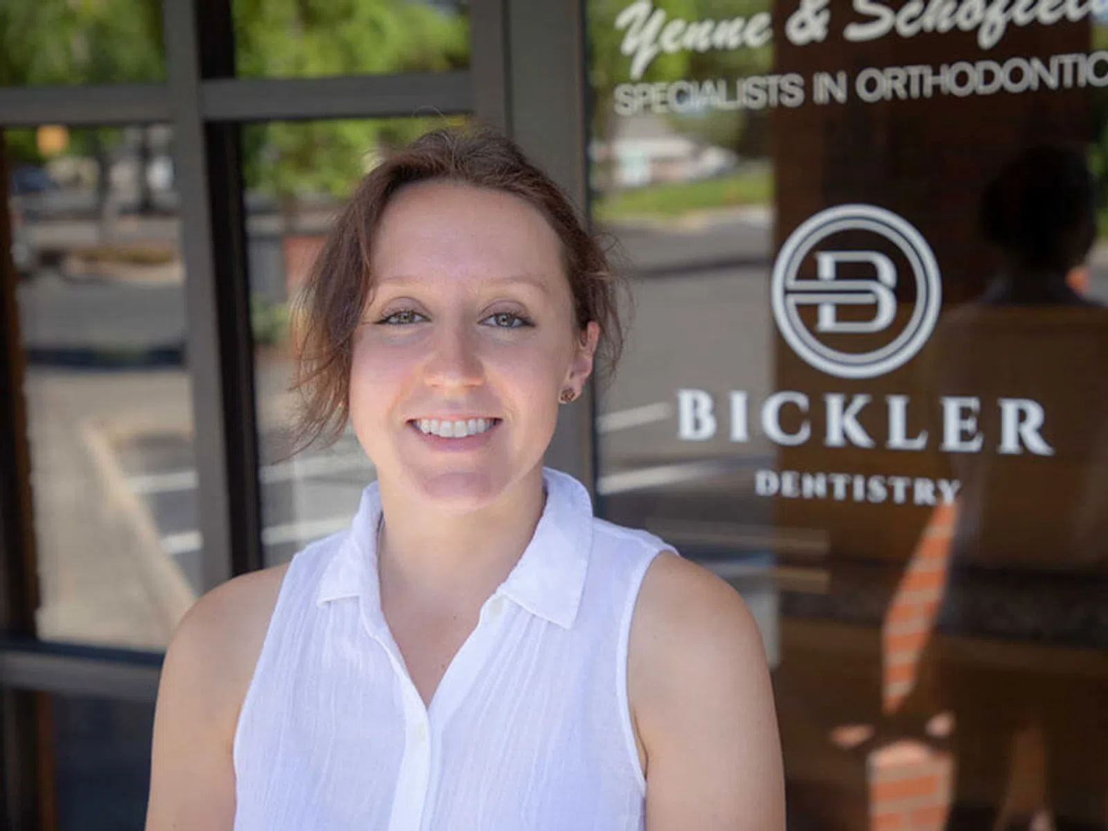 Smiling woman in white sleeveless blouse standing in front of Bickler Dentistry and orthodontics office door.