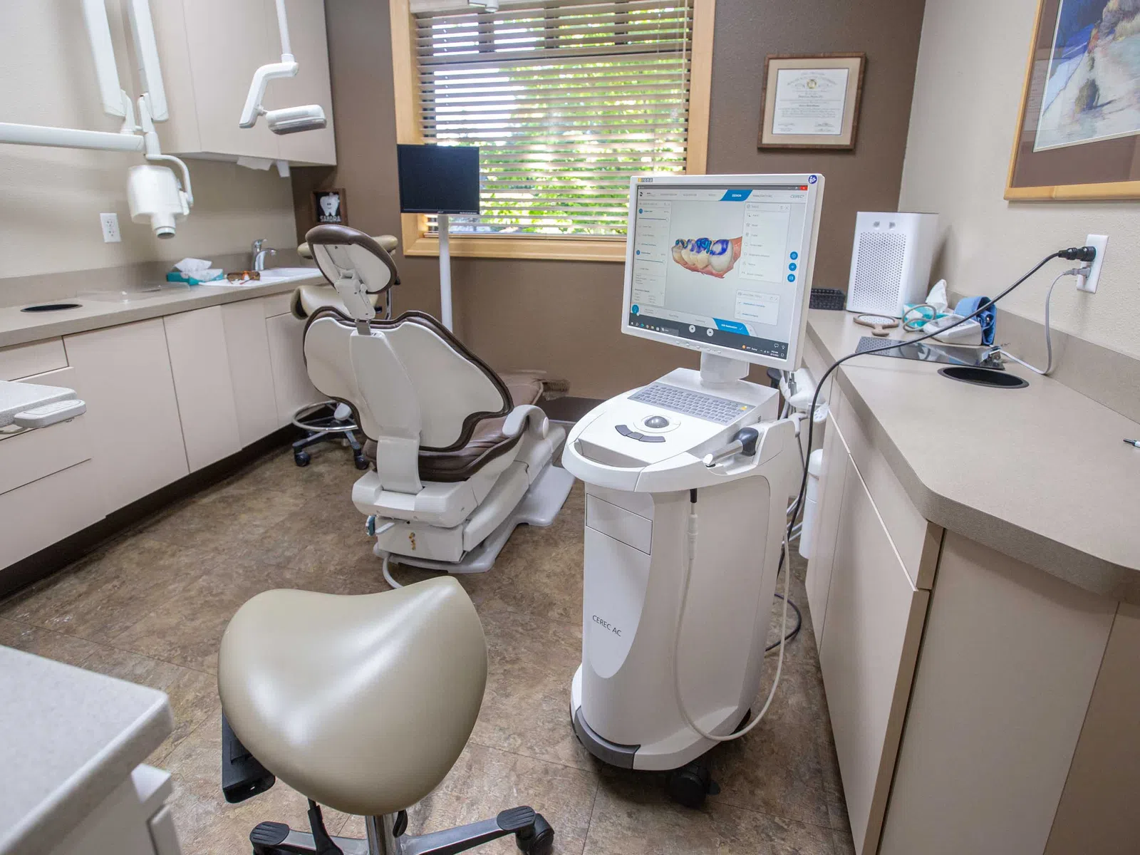 Modern dental office with dental chair, imaging equipment, and computer displaying dental scan near window.