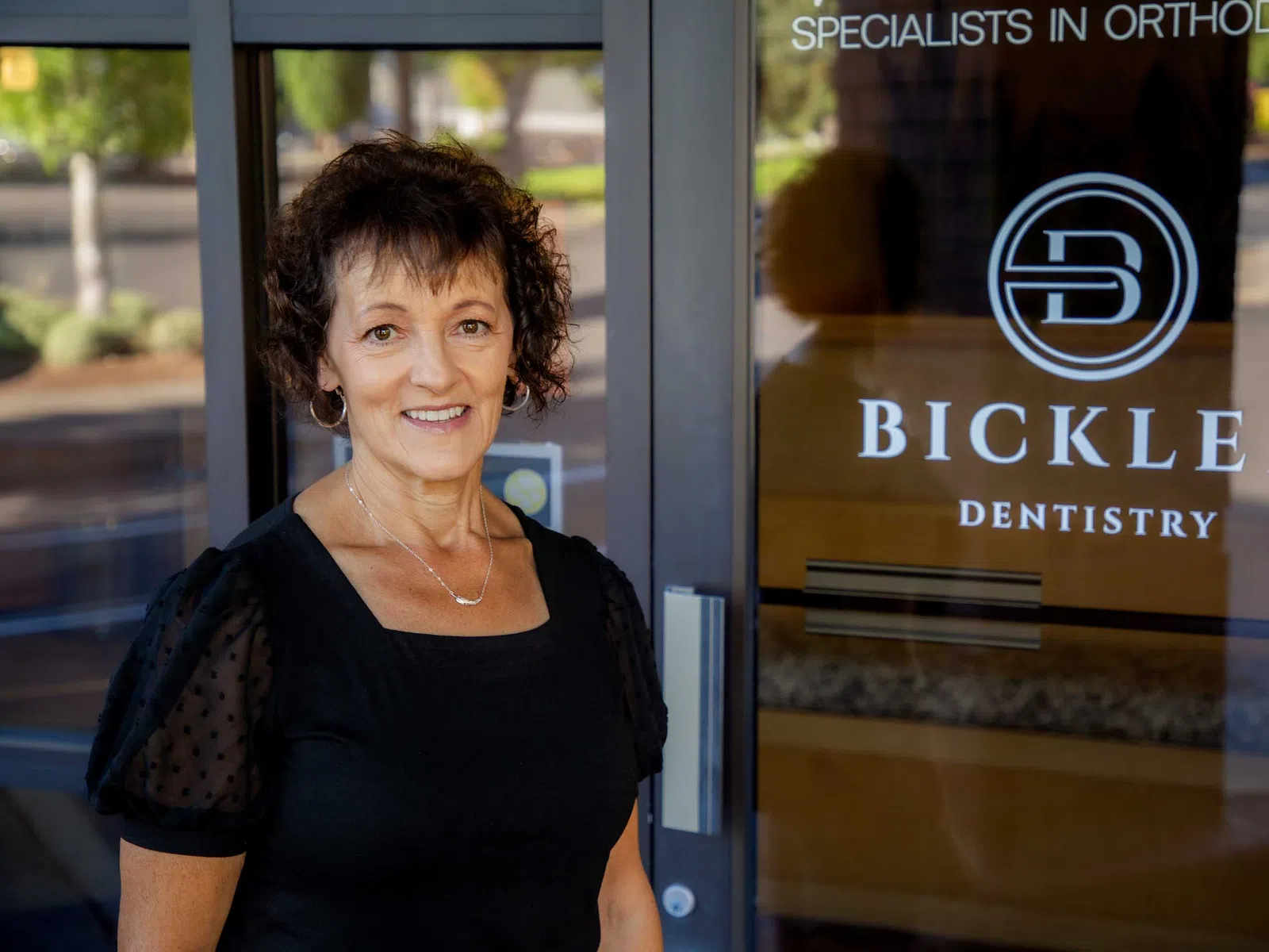 Smiling woman standing outside Bickler Dentistry clinic with visible logo on glass door under daylight.