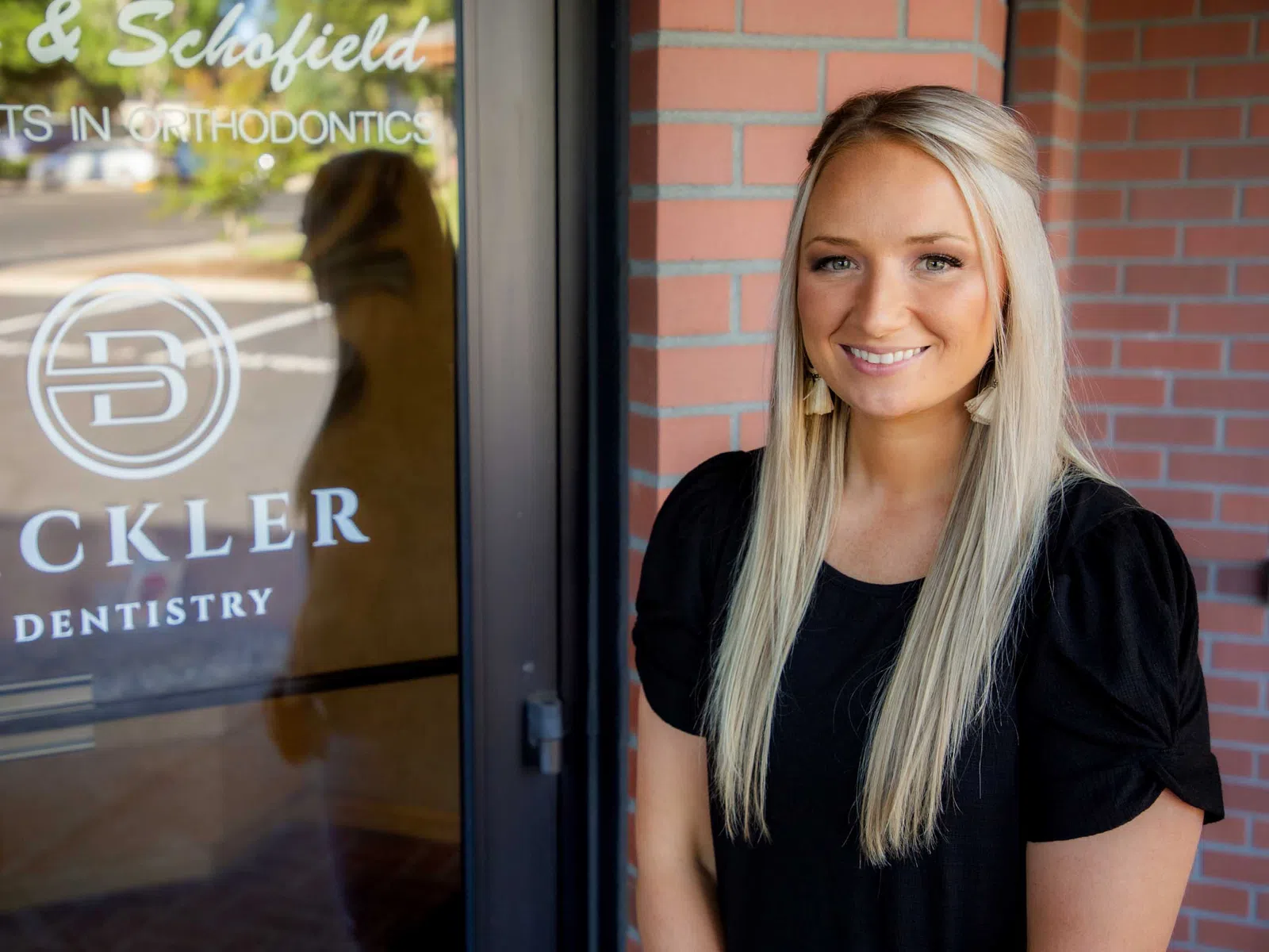 Smiling woman with long blonde hair stands outside Bickler Dentistry and Orthodontics office near brick wall.