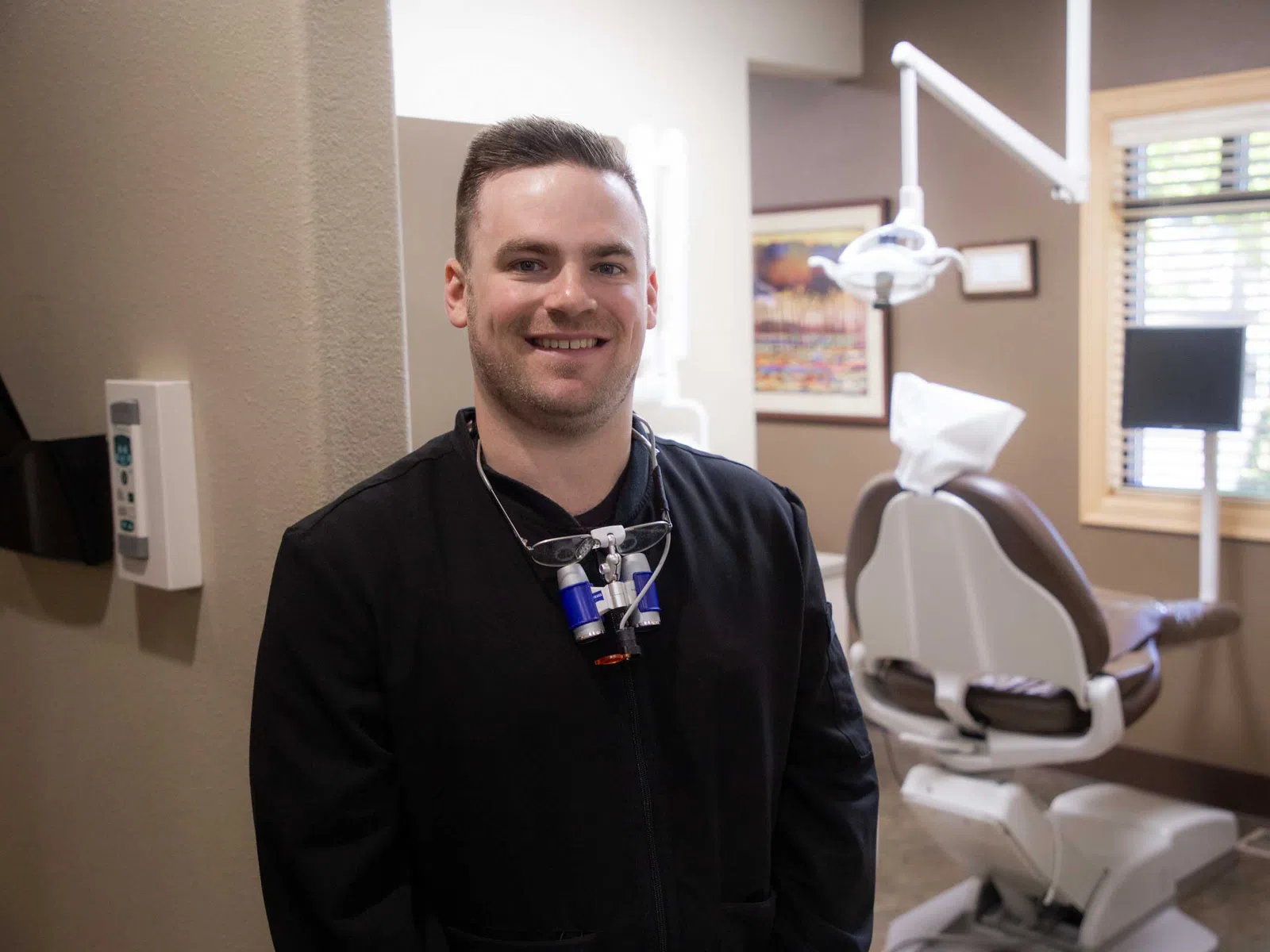 Smiling male dentist standing in a modern dental office with equipment and patient chair behind him.