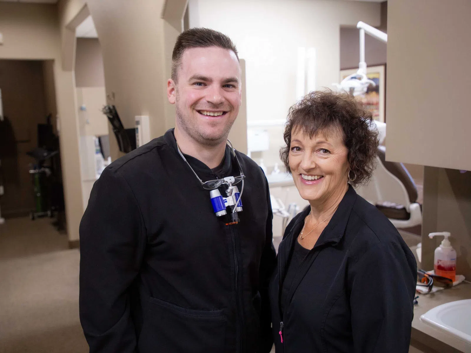 Smiling male dentist and female dental assistant in black uniforms standing in a dental clinic hallway.