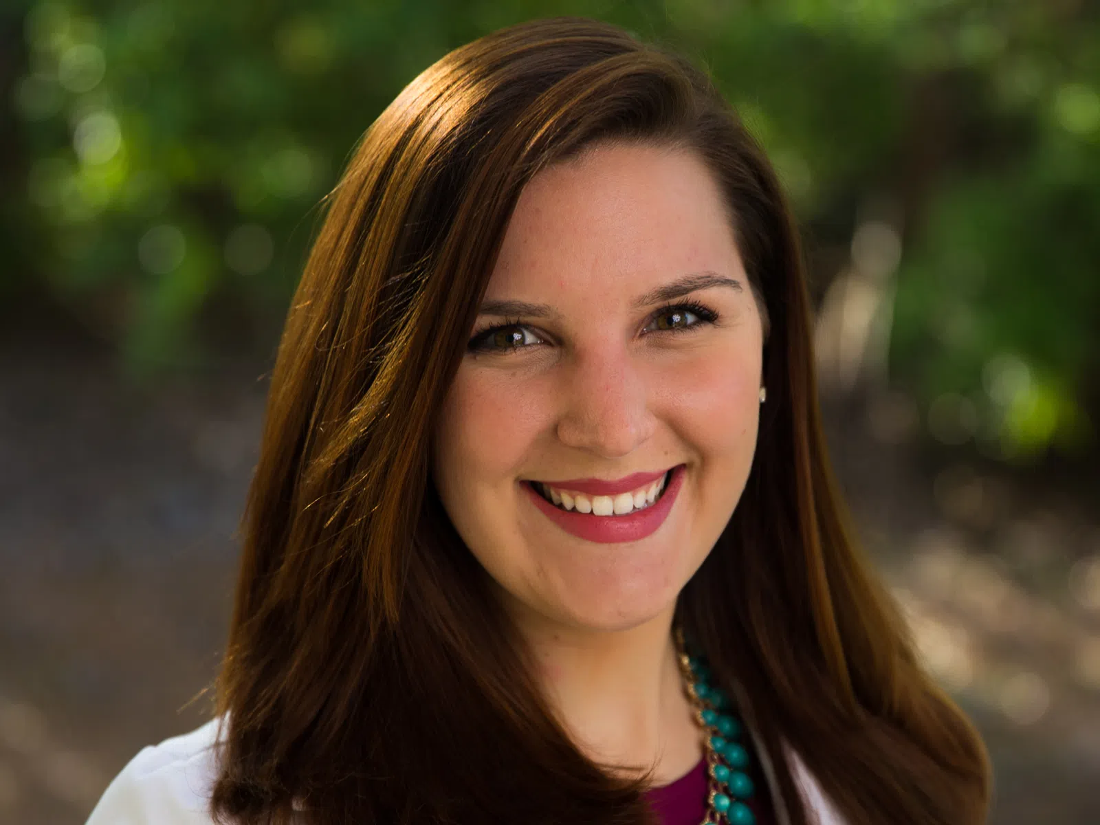 Smiling woman with long brown hair wearing a white coat and turquoise necklace outdoors in natural light
