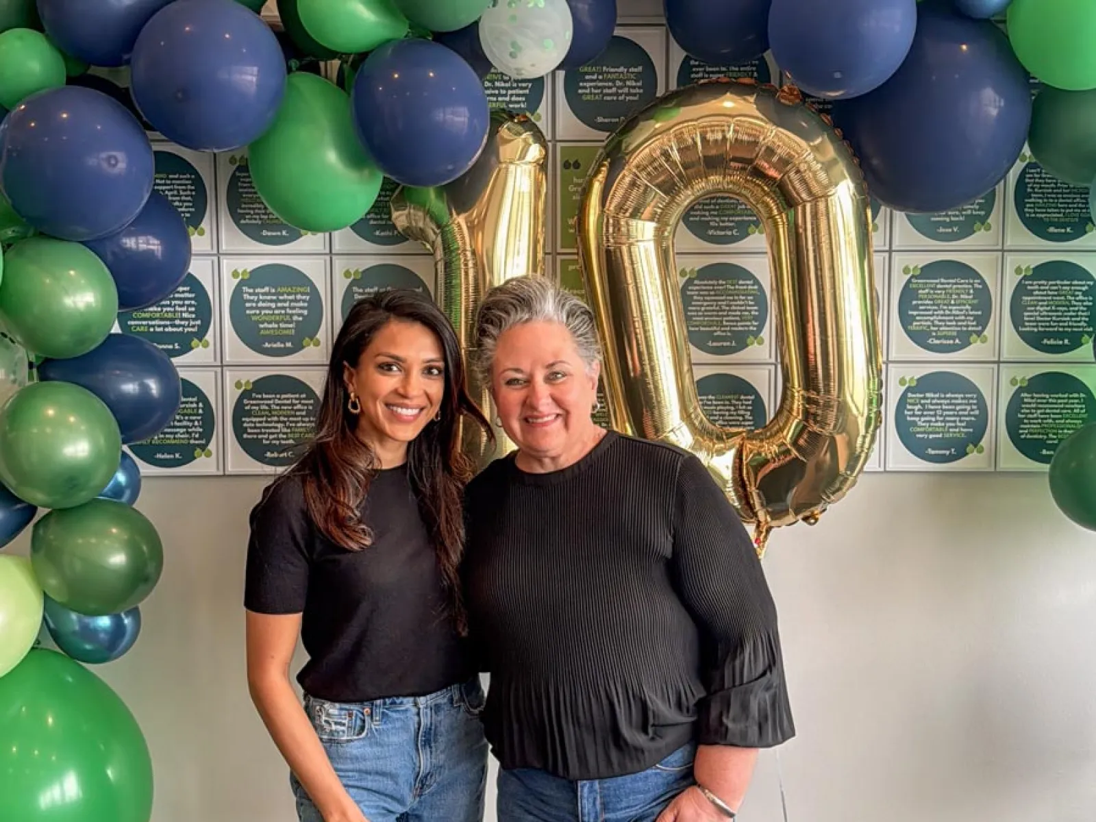 Two women smile together in front of gold '10' balloons and green and blue balloon arch celebrating milestone.