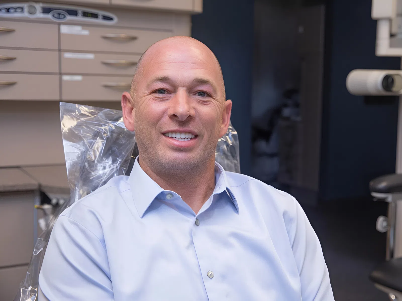 Smiling bald man in a blue dress shirt sitting in a dental office chair with cabinets in the background