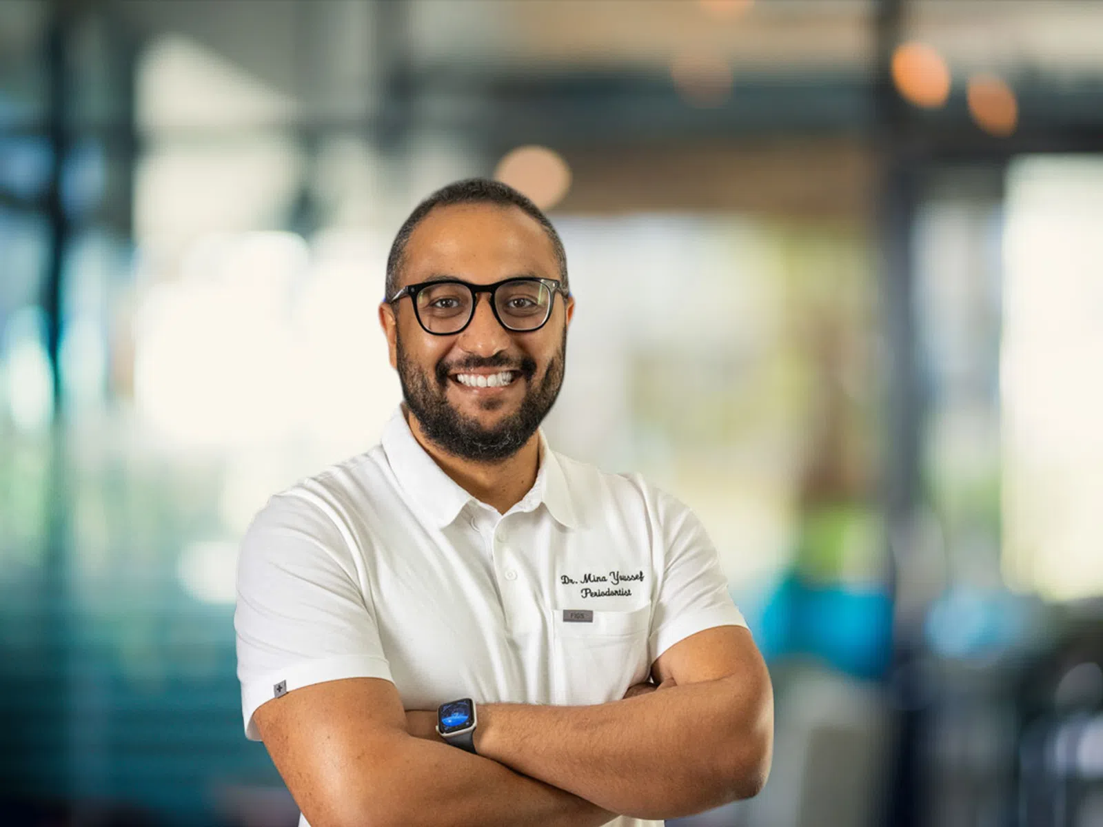 Smiling man with glasses and beard wearing a white shirt crosses arms in a modern office environment.