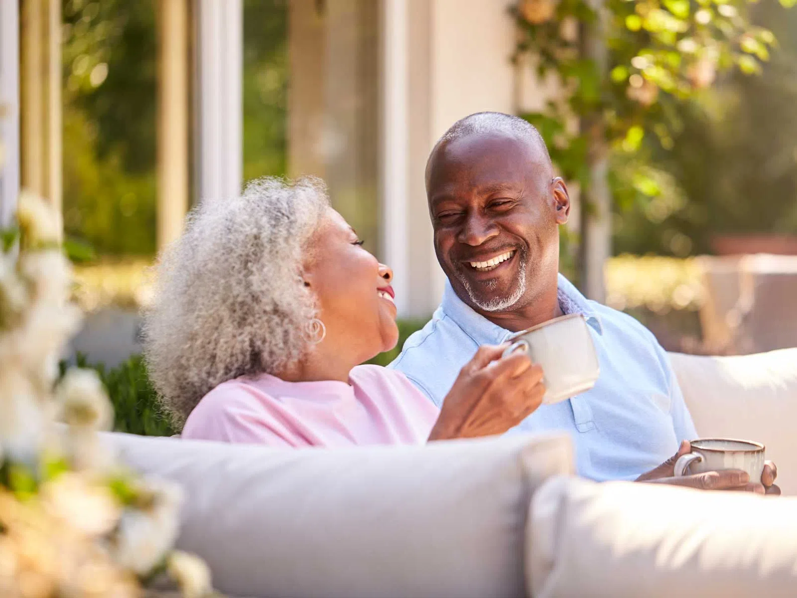 Older couple sitting outdoors on a sofa, smiling and enjoying coffee together on a sunny day.