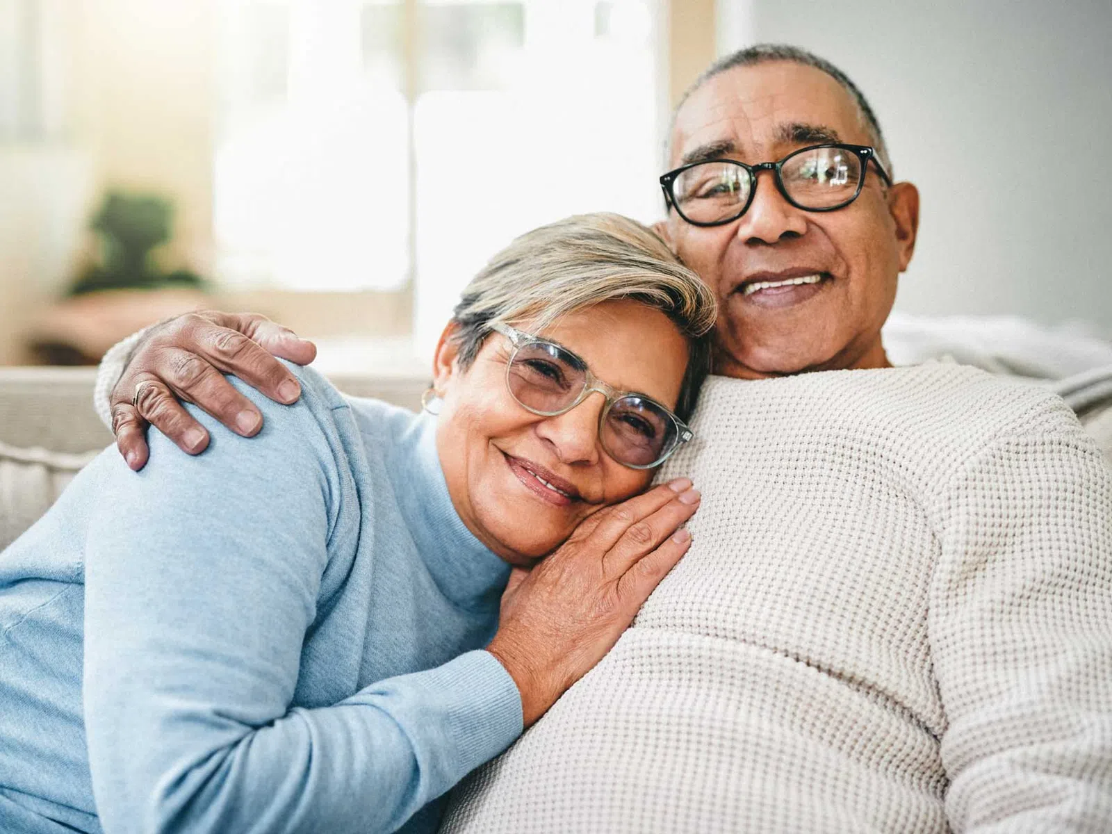 Smiling elderly couple embracing on a couch in a cozy living room with natural light.