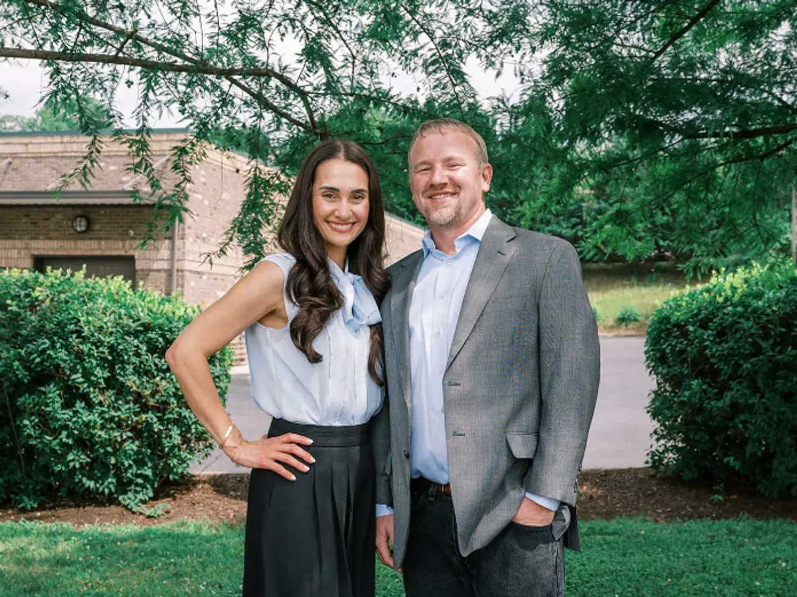Smiling man and woman dressed professionally standing outside near green bushes and trees on a sunny day.