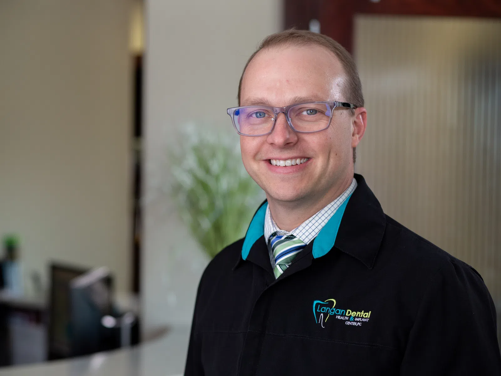 Smiling male dentist wearing glasses and Langan Dental jacket standing in dental office with blurred background.