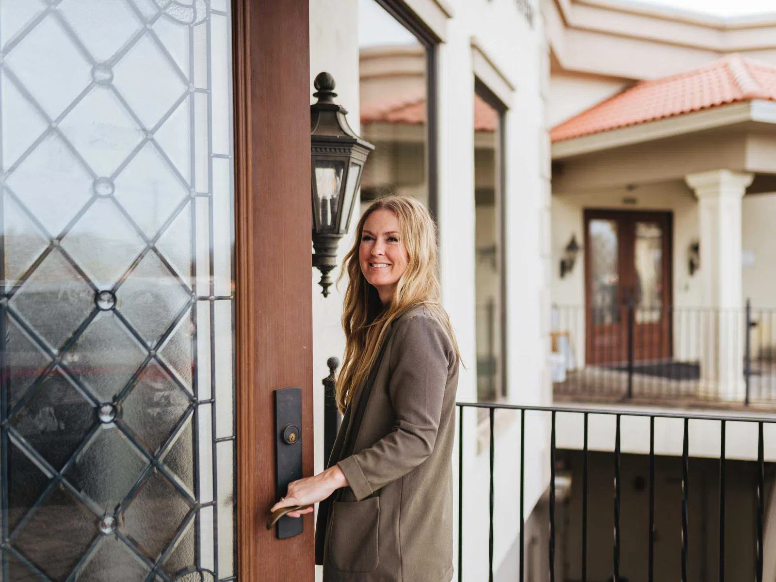 Smiling woman in brown jacket opening a wooden door with decorative glass on a residential porch