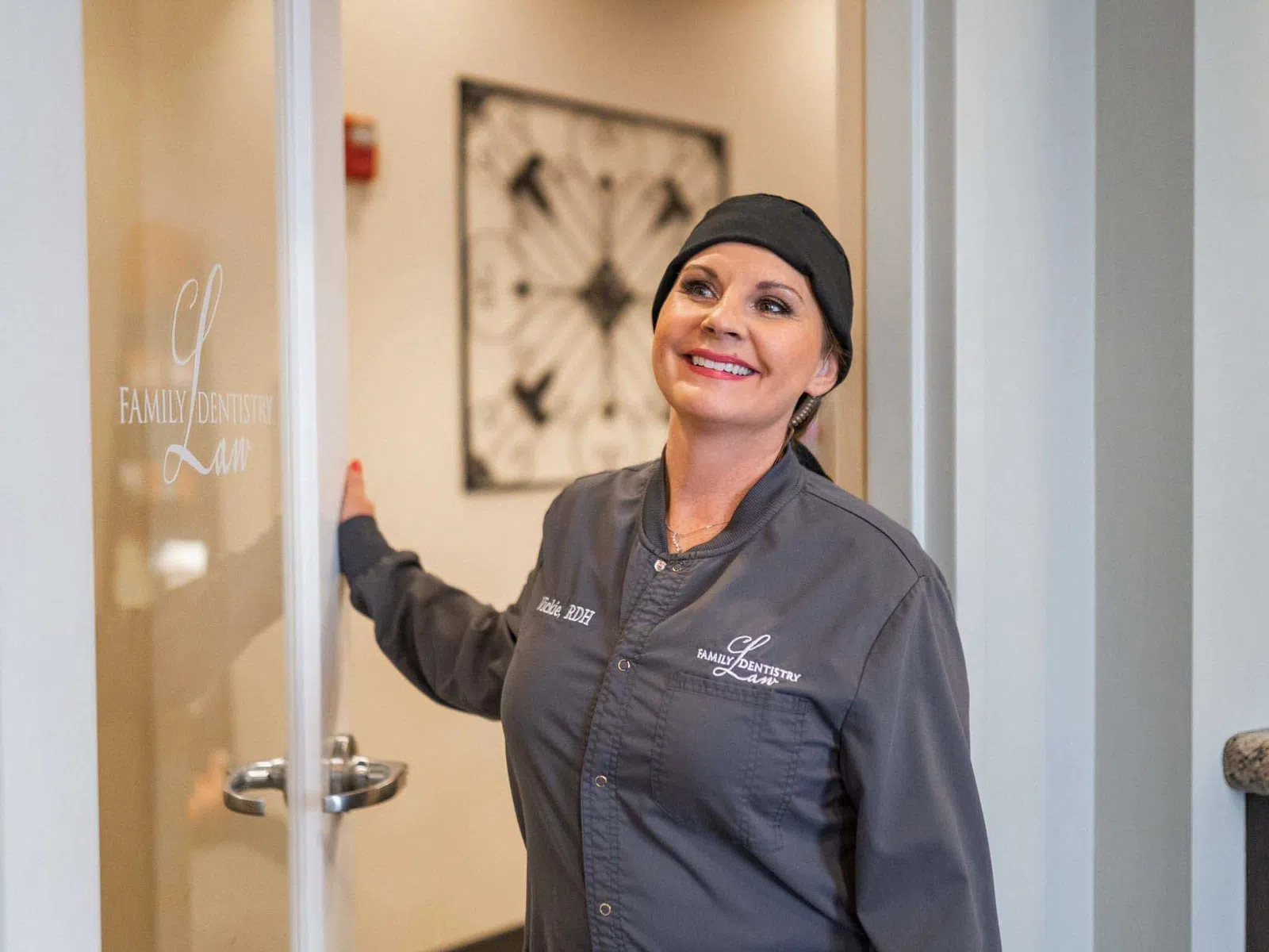 Smiling female dentist in uniform holding open a glass door with Family Dentistry logo in a clinic.