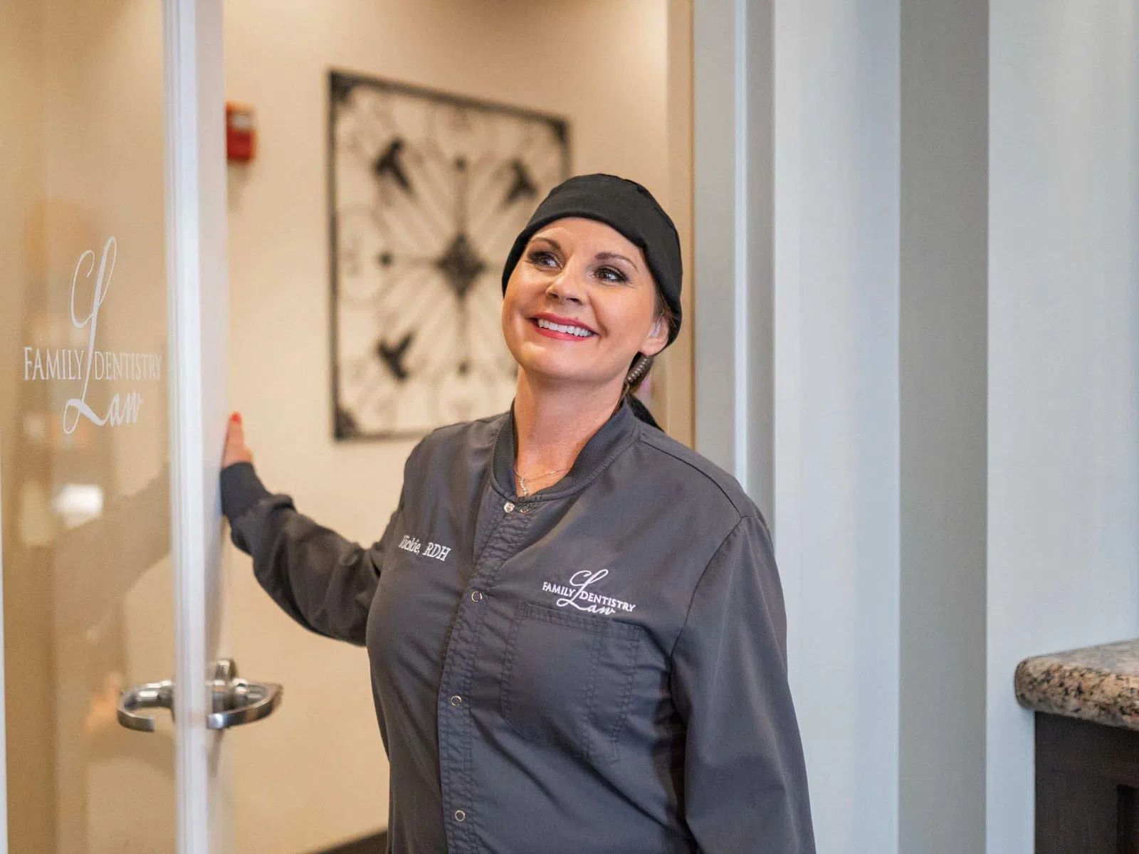Smiling female dental professional in gray uniform opening door at a modern family dentistry clinic.