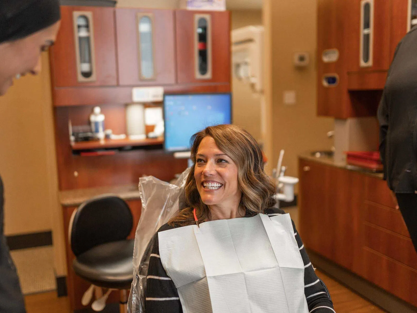 Smiling woman sitting in dental chair with bib talking to a dentist in a modern dental office.