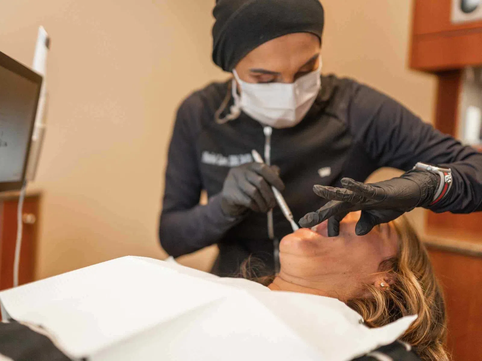 Dentist wearing mask and gloves treating a female patient reclining in a dental chair with equipment in view