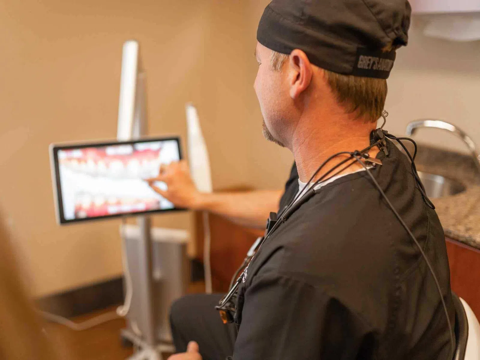 Dentist in scrubs pointing at dental x-ray images on a screen during patient consultation.