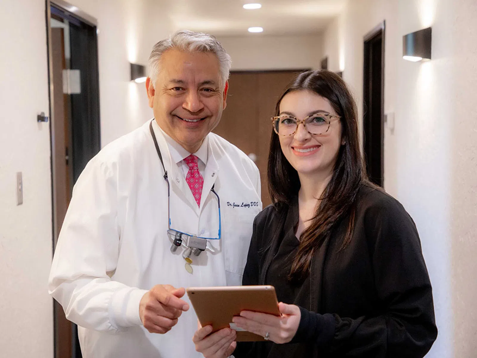 Smiling male dentist and female patient holding a digital tablet in a clinic hallway.
