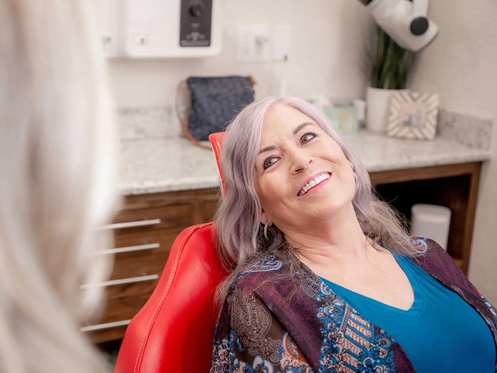 Smiling older woman with gray hair sitting in a red chair in a bright modern medical office.