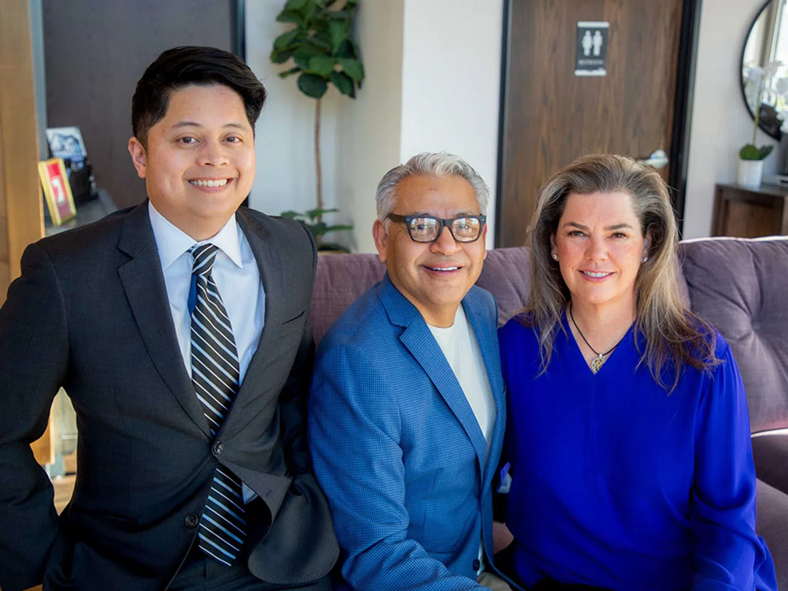 Three professionally dressed people smiling indoors on a couch with modern decor and natural light.